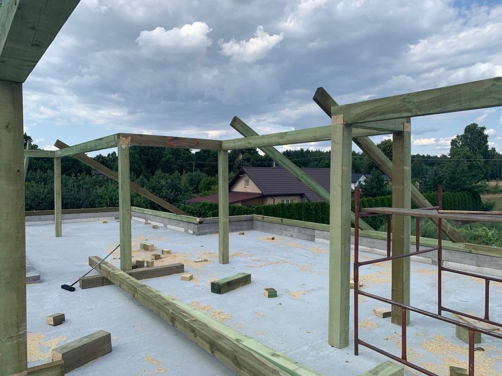 Konstrukcja drewniana dachu w trakcie budowy, widoczny szkielet z impregnowanego drewna na betonowej płycie, w tle dom jednorodzinny i zieleń.