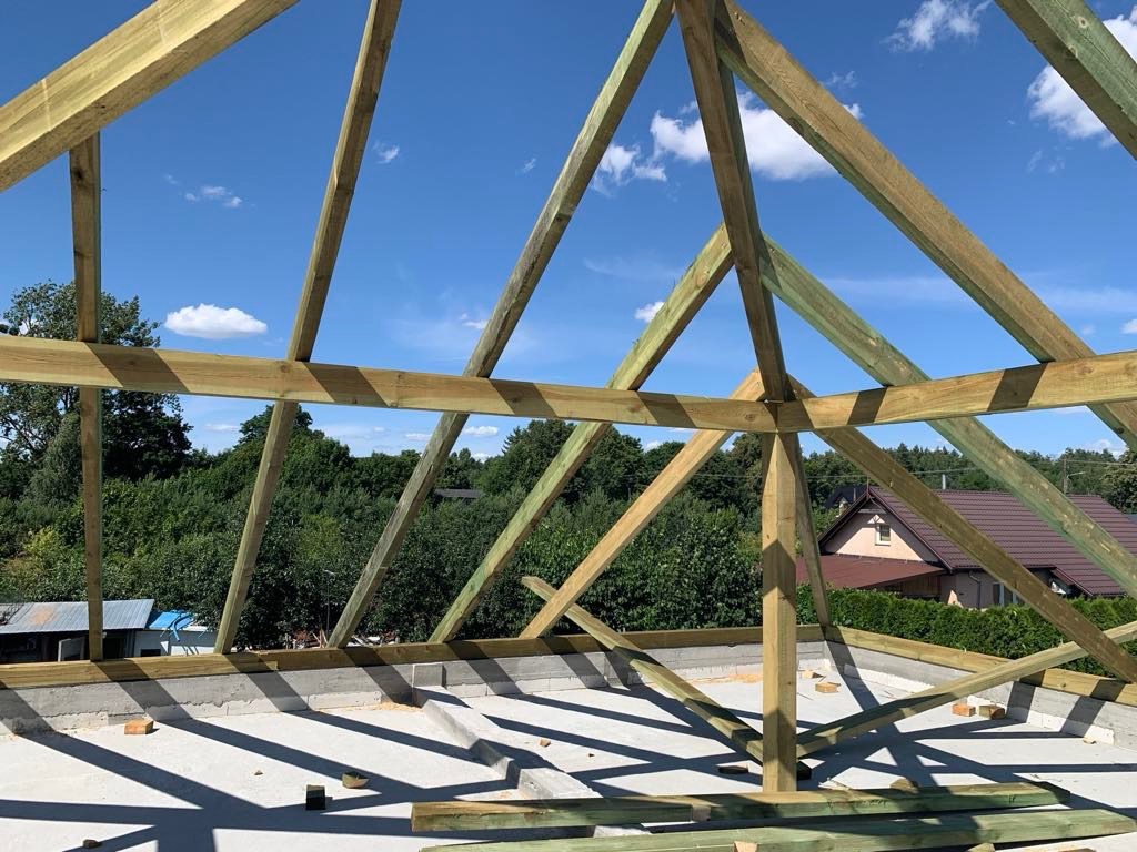 Konstrukcja drewniana dachu w trakcie budowy domu w Warszawie, widok z góry na betonowy strop i elementy więźby dachowej na tle zieleni.