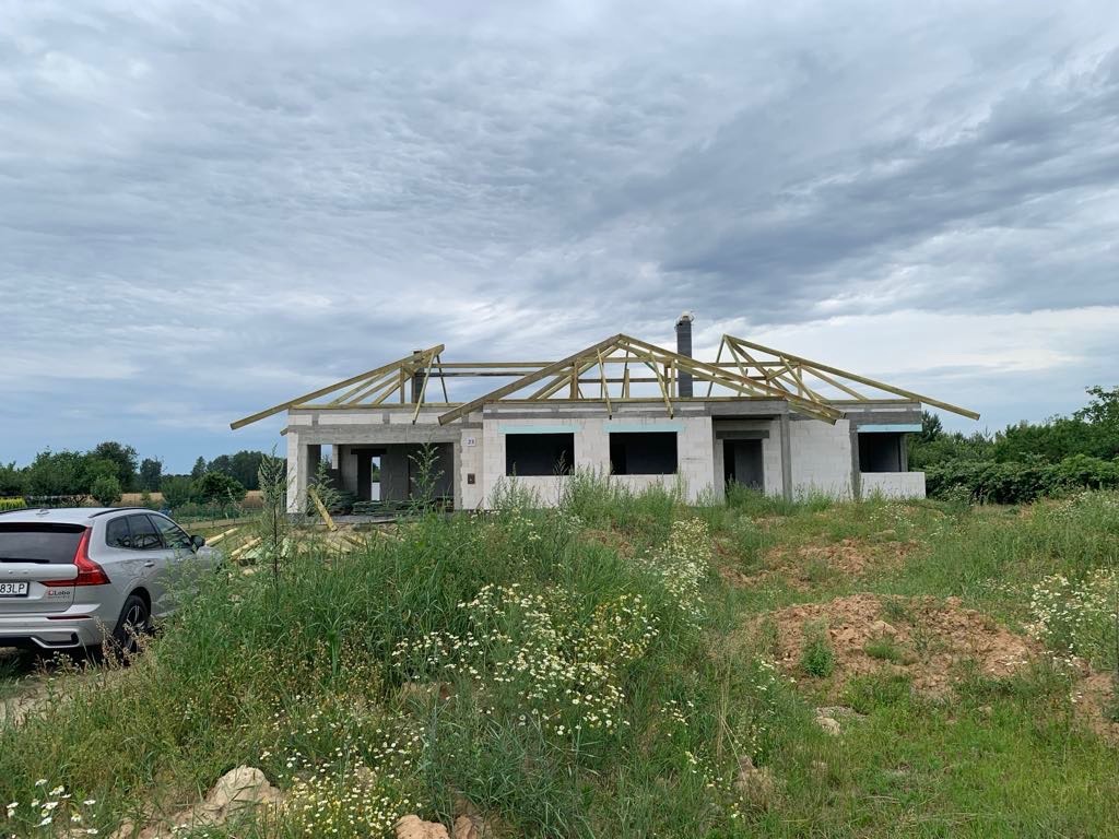 Surowy stan domu jednorodzinnego z konstrukcją dachu w trakcie budowy, widok z poziomu gruntu, zarośnięte pole na pierwszym planie, szary samochód osobowy zaparkowany obok.