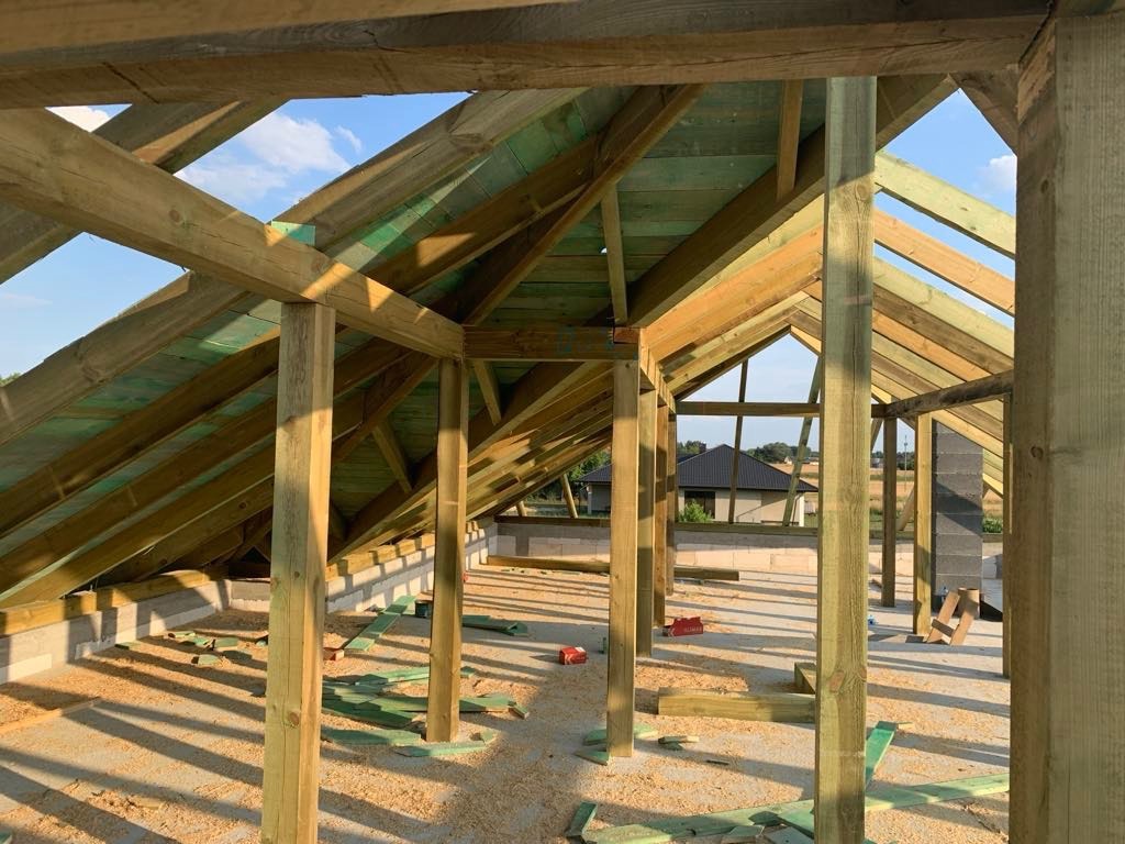 Konstrukcja drewniana dachu w trakcie budowy domu murowanego, widoczne bale nośne, deski poszycia, bloczki betonowe ścian i komin z cegły klinkierowej w tle krajobraz wiejski