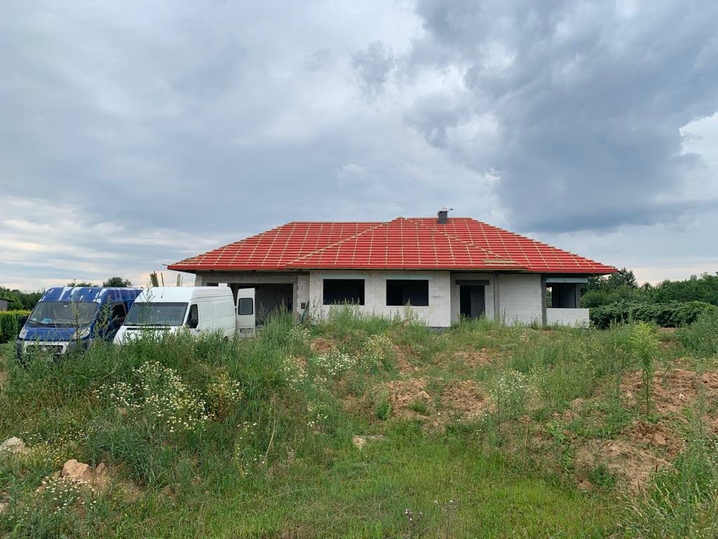 Surowy stan domu jednorodzinnego z czerwoną dachówką w trakcie budowy, widoczne dwa busy dostawcze na pierwszym planie, zachmurzone niebo.