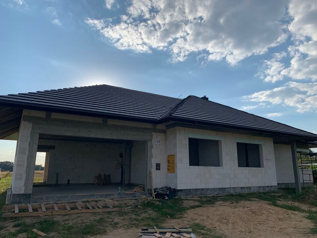 Surowy stan domu jednorodzinnego z ciemną dachówką na tle błękitnego nieba z chmurami, widoczne pustaki i otwory okienne bez ram.