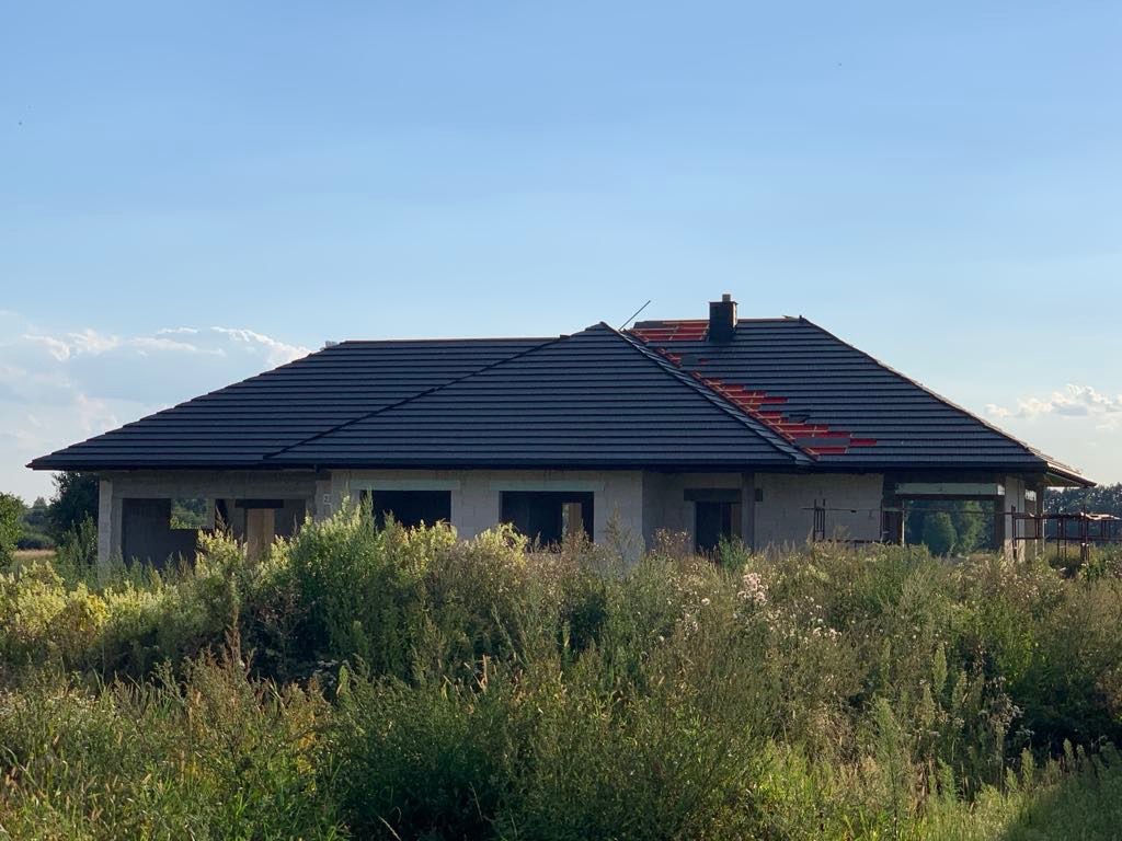 Surowy budynek jednorodzinny z ciemnym dachem w trakcie budowy, widok z poziomu wysokiej trawy, częściowo położona dachówka.