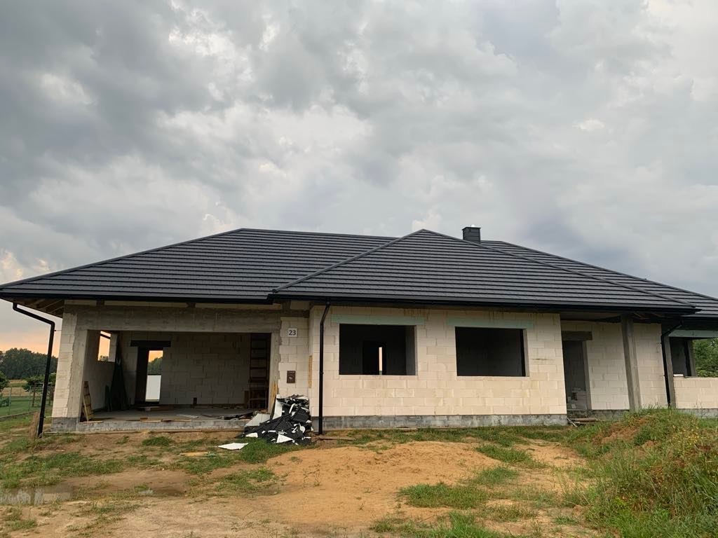 Surowy budynek z białych pustaków z czarnym dachem z dachówki, widoczny numer domu '23', etap budowy w trakcie prac wykończeniowych, ponure niebo w tle.
