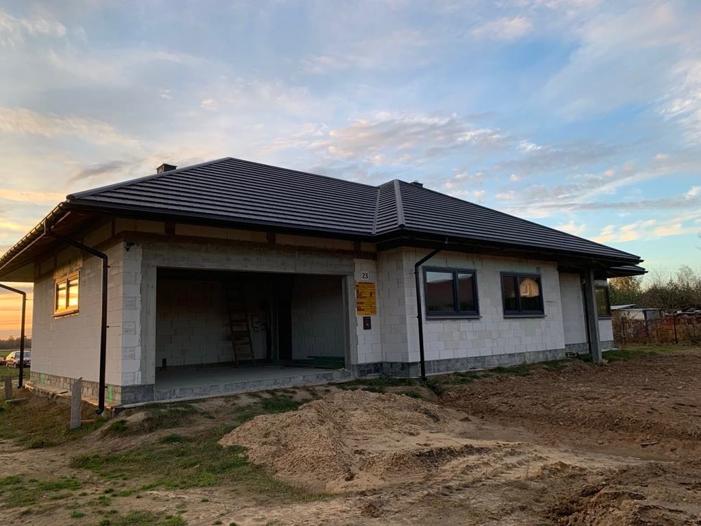 Niedokończony dom murowany z garażem w stanie surowym otwartym, widoczne pustaki, czarna dachówka i numer domu 23, ziemia wokół budynku nierówna.