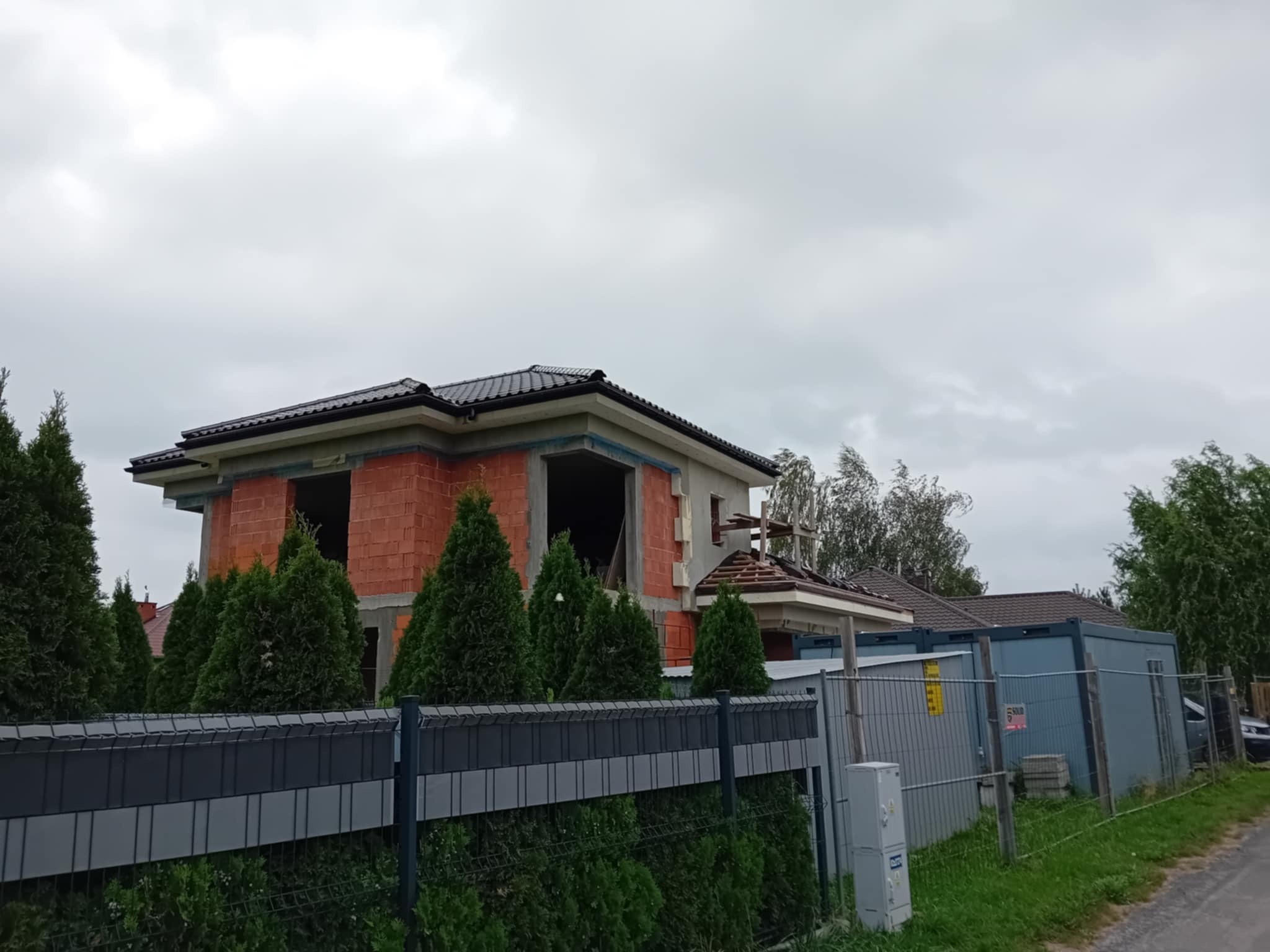 Surowy stan domu z czerwonej cegły pod ciemnym dachem, widoczny zza ogrodzenia i żywopłotu z tuj, z kontenerem w tle.