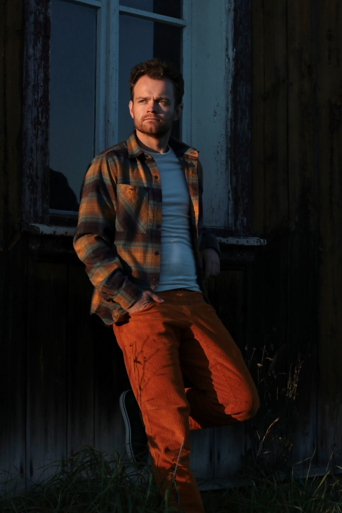 Młody mężczyzna w flanelowej koszuli i pomarańczowych spodniach sztruksowych oparty o drewniany budynek z oknem, oświetlony zachodzącym słońcem.