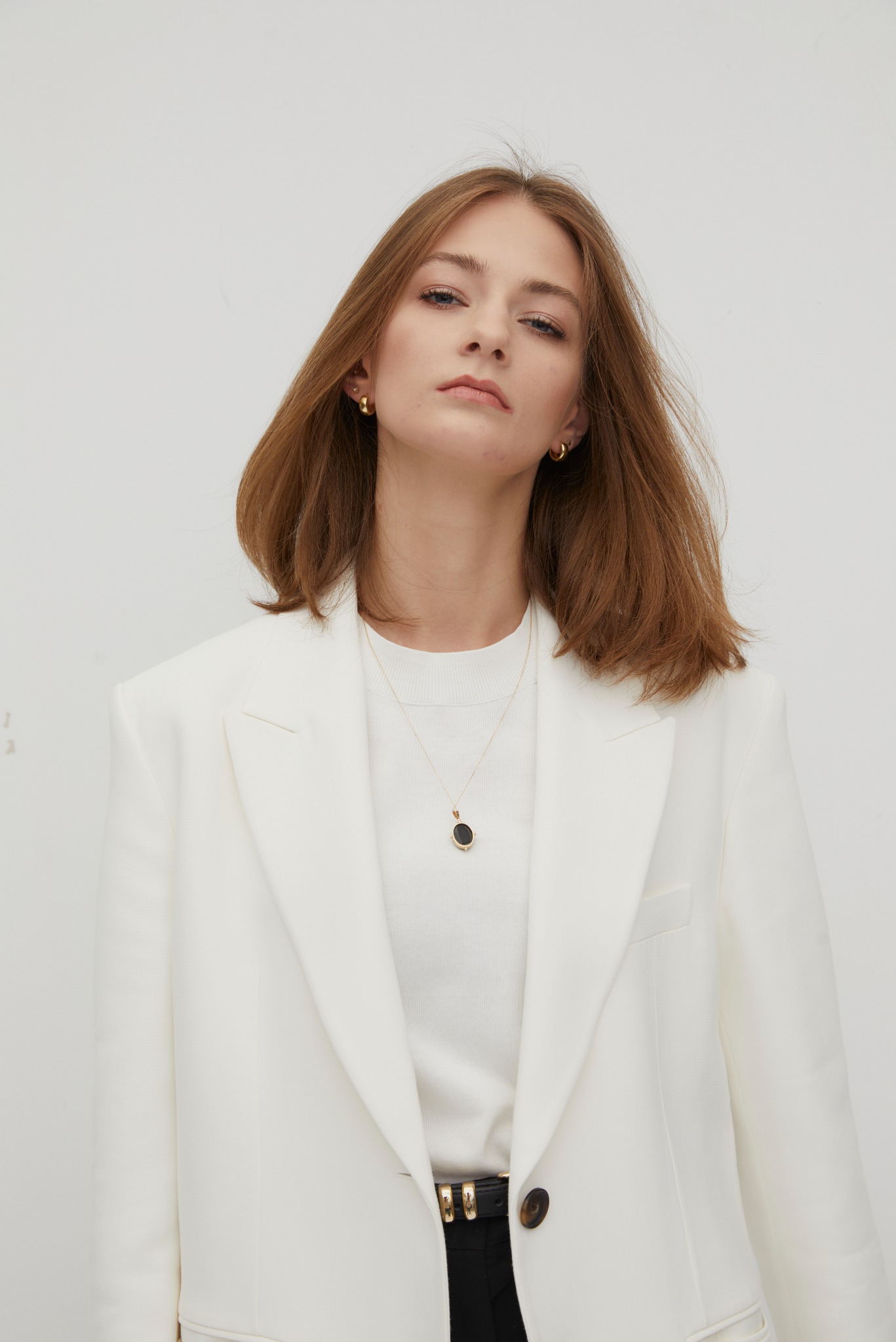 Portret kobiety w białym garniturze, z delikatnym makijażem i złotą biżuterią na tle jasnej ściany.