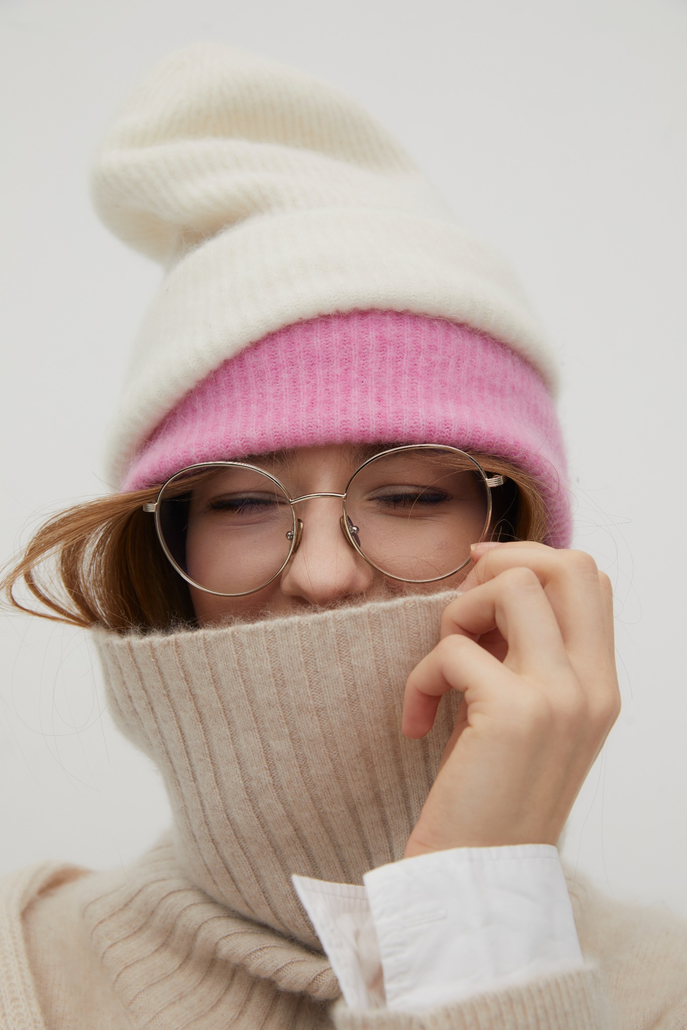 Portret kobiety w okularach, z twarzą częściowo zakrytą beżowym golfem, w dwukolorowej czapce (biało-różowej), z rudymi włosami wystającymi spod czapki.