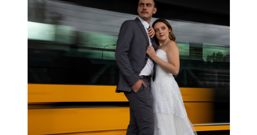 Para młoda pozuje na tle pędzącego tramwaju, mężczyzna w szarym garniturze z ręką w kieszeni, kobieta w białej sukni ślubnej opiera się o jego ramię.