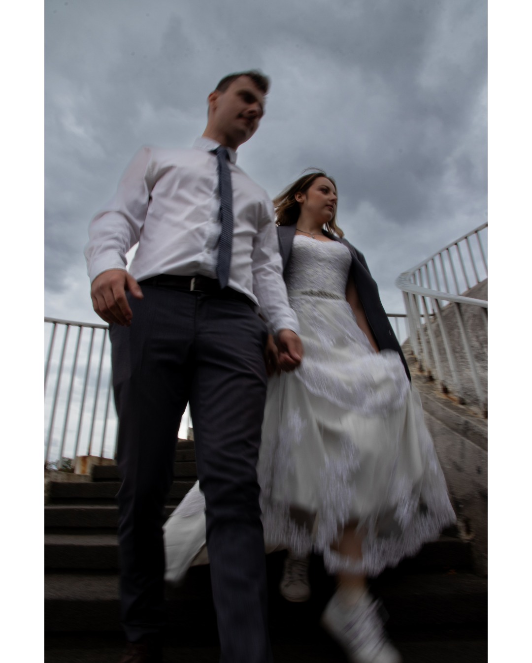 Para młoda schodzi po schodach trzymając się za ręce, pochmurne niebo w tle, panna młoda w sukni ślubnej i sportowych butach.