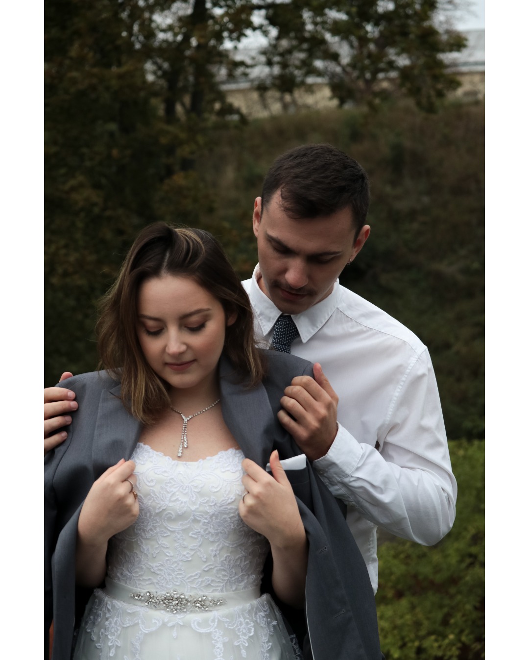 Panna młoda w koronkowej sukni ślubnej, z szarą marynarką na ramionach, spogląda w dół, a pan młody w białej koszuli i krawacie pomaga jej założyć marynarkę, na tle zieleni.