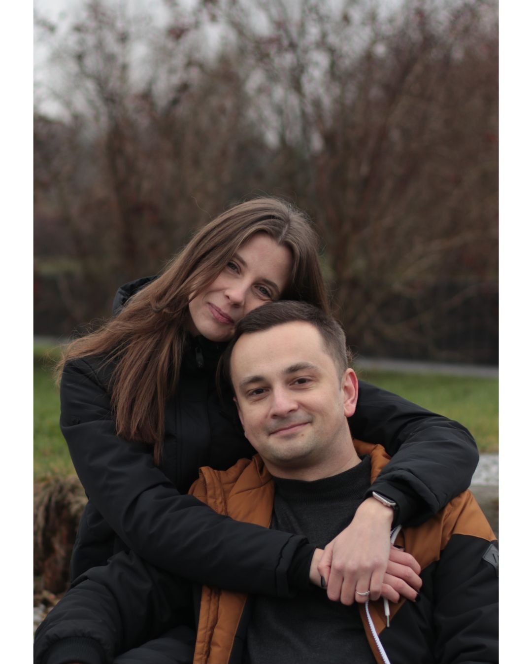 Para obejmująca się na zewnątrz w pochmurny dzień, kobieta z długimi brązowymi włosami w czarnej kurtce obejmuje mężczyznę w brązowej kamizelce, oboje patrzą w obiektyw z uśmiechem.