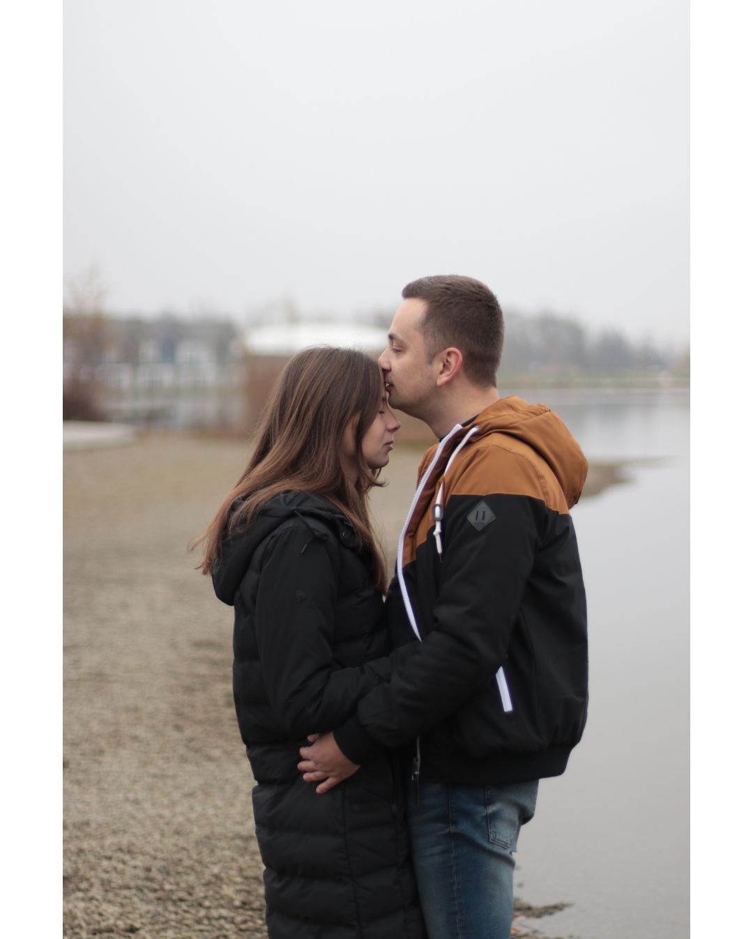 Para zakochanych przytulona na tle jeziora w pochmurny dzień, mężczyzna całuje kobietę w czoło.
