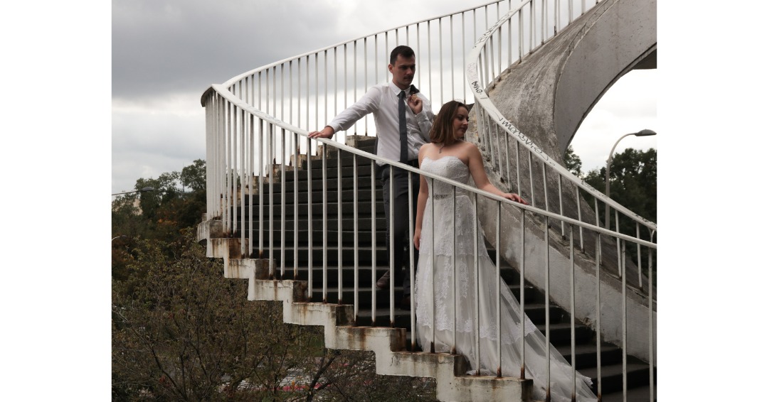 Para młoda schodzi po spiralnych, betonowych schodach na tle pochmurnego nieba; panna młoda w koronkowej sukni bez ramiączek, pan młody w jasnej koszuli z marynarką przewieszoną przez ramię.