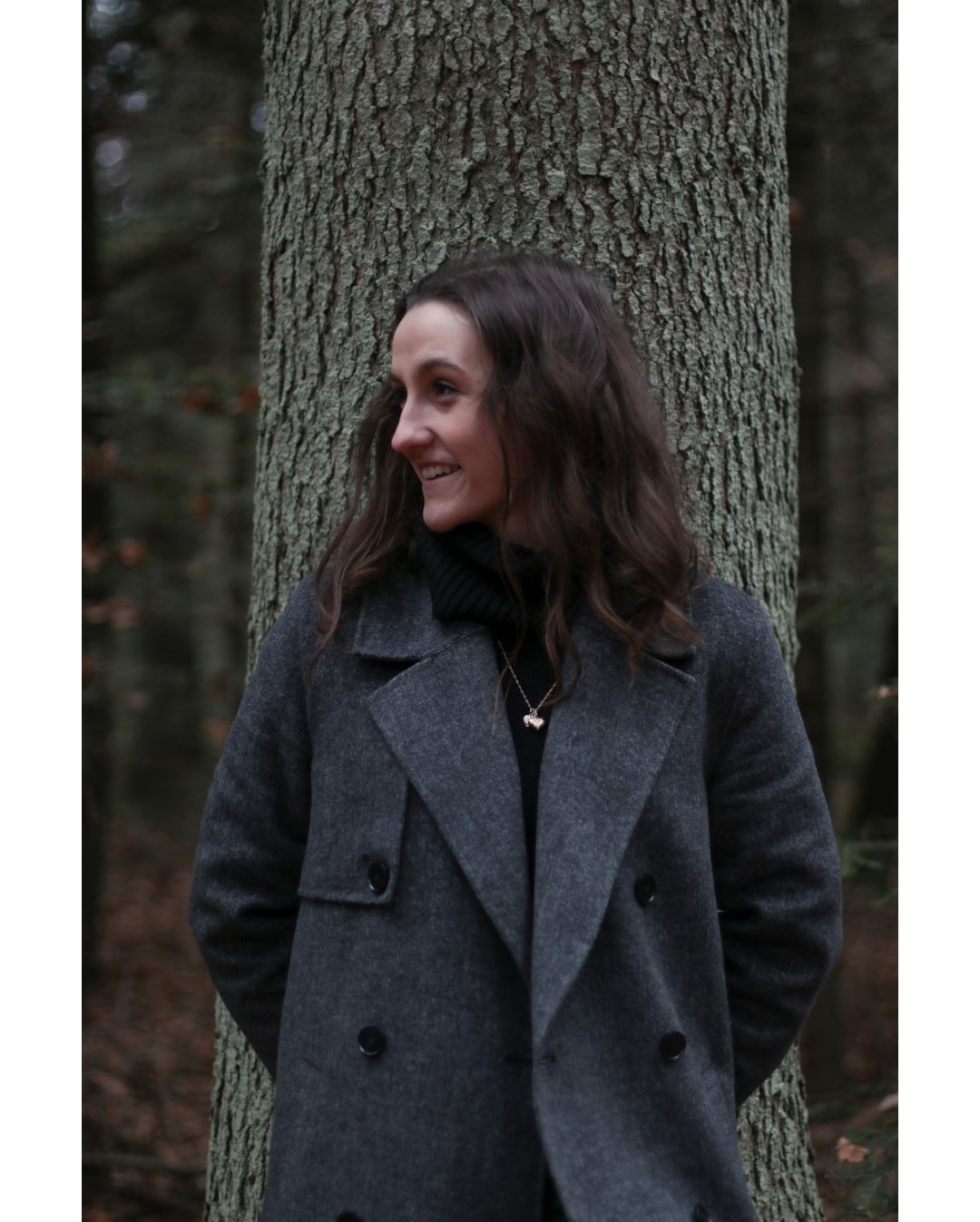 Uśmiechnięta kobieta w szarym płaszczu opiera się o drzewo na tle leśnym, widok z profilu.