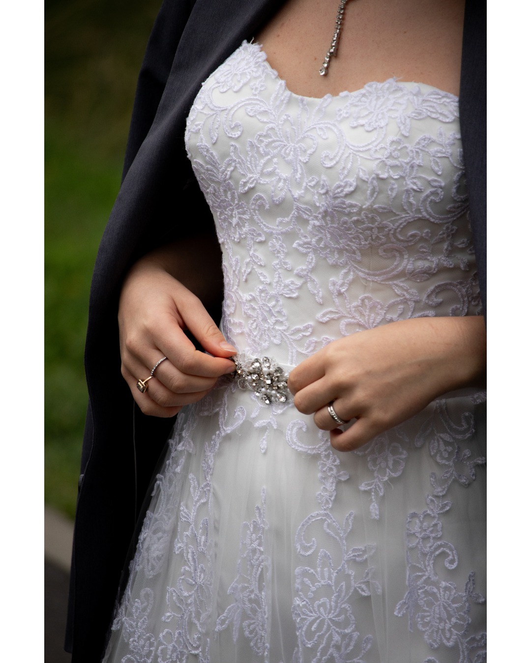 Panna młoda zapinająca ozdobny, srebrny pasek z kryształkami do koronkowej sukni ślubnej, marynarka na ramionach.