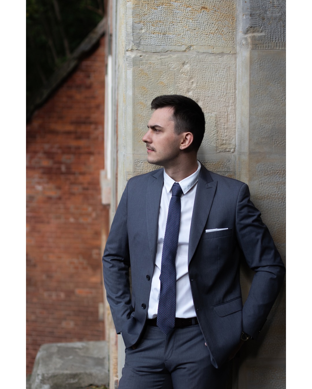 Mężczyzna w szarym garniturze, białej koszuli i granatowym krawacie w kropki, oparty o kamienną ścianę, patrzy w lewo. W tle mur z czerwonej cegły.