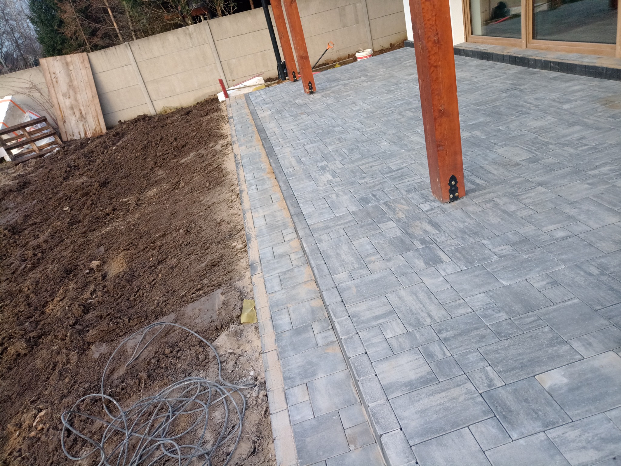 Świeżo ułożona szara kostka brukowa na posesji z drewnianymi słupami, widoczna ziemia i betonowe ogrodzenie w tle.