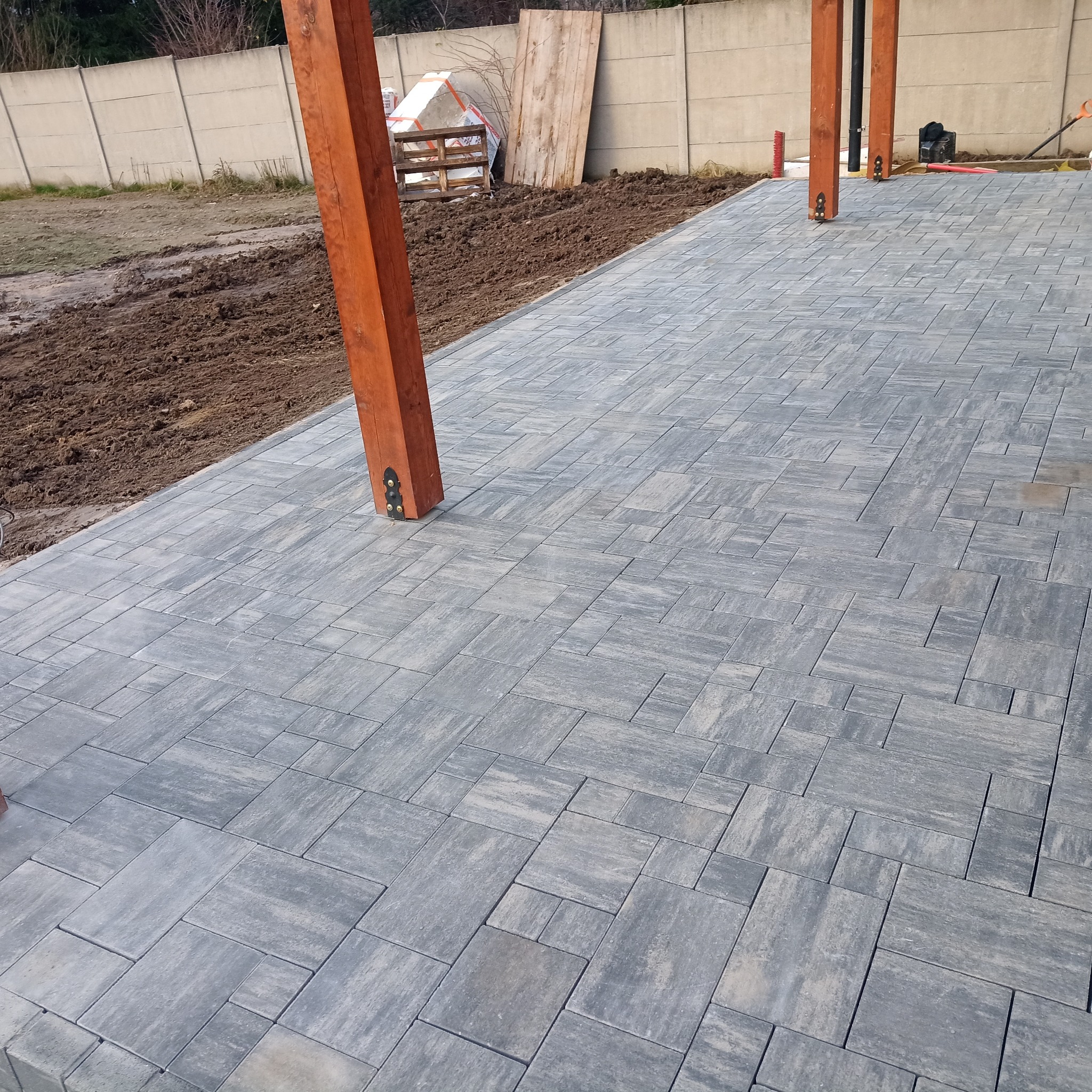 Ułożona szara kostka brukowa tworząca wzór na podwórku, z drewnianymi słupami podtrzymującymi konstrukcję i widocznym fragmentem przygotowanego terenu pod dalsze prace.