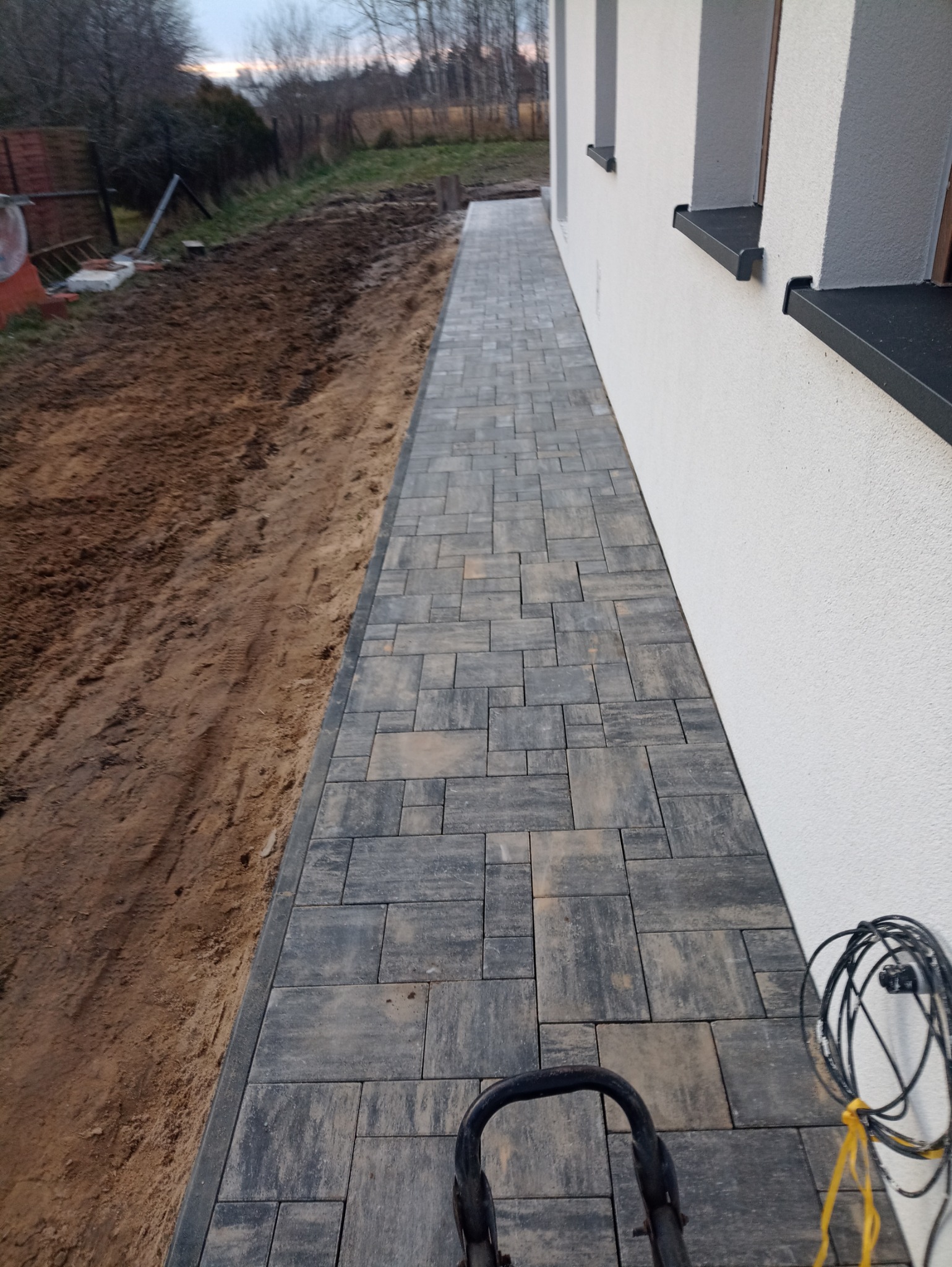 Ułożona szara kostka brukowa tworząca chodnik wzdłuż białej ściany budynku, z widocznym obrzeżem i przygotowanym terenem po drugiej stronie, częściowo zasłonięta ubijarką w dolnej części kadru...