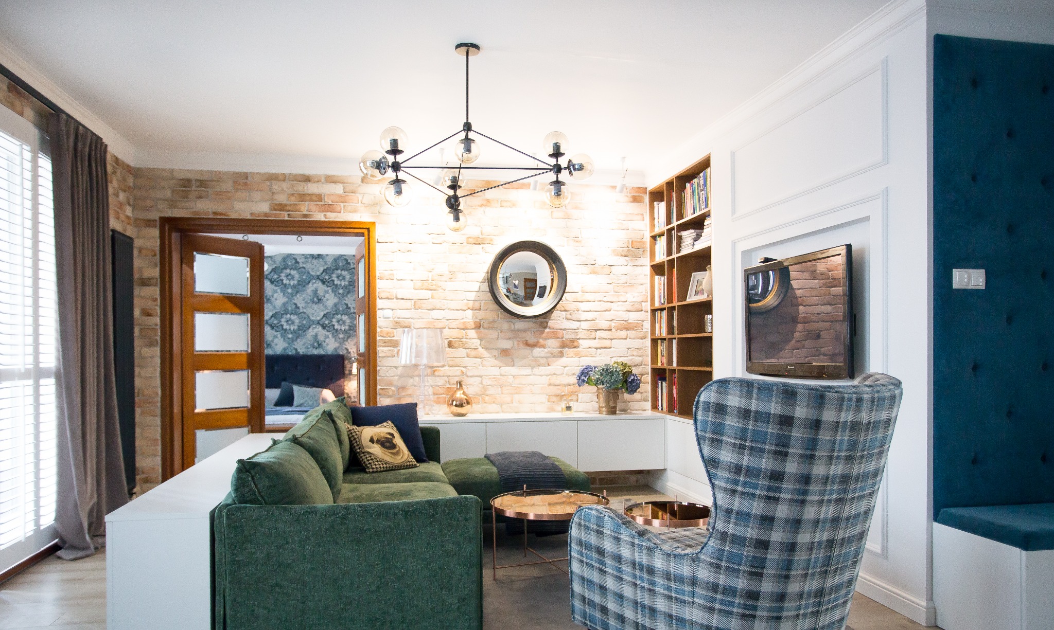 Salon z ceglaną ścianą, zieloną sofą, wzorzystym fotelem i nowoczesnym oświetleniem, widok na sypialnię z tapetą w tle.
