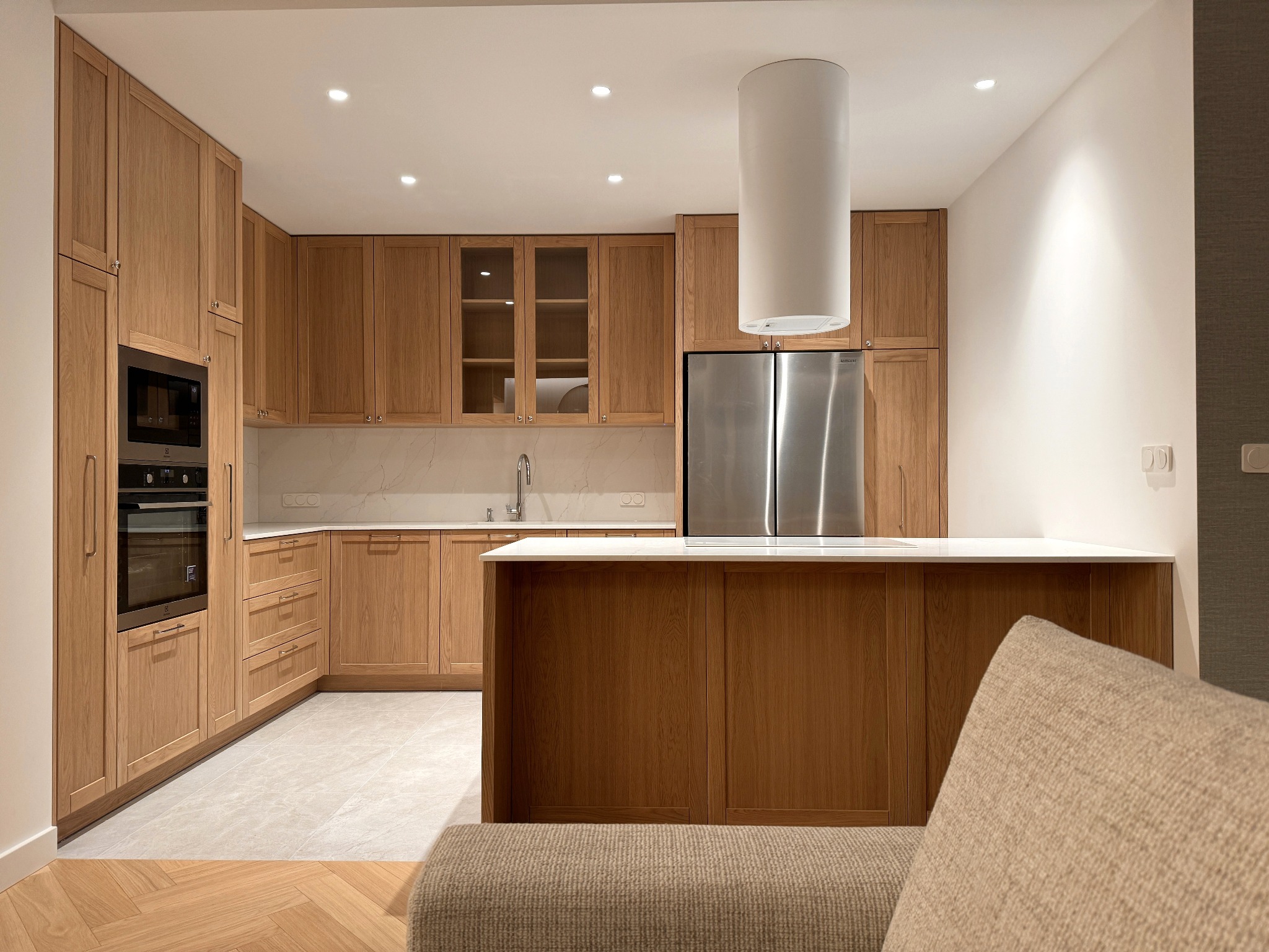 Jasna kuchnia z drewnianymi szafkami, białym blatem i wyspą kuchenną, widok z perspektywy salonu z fragmentem sofy w kadrze.