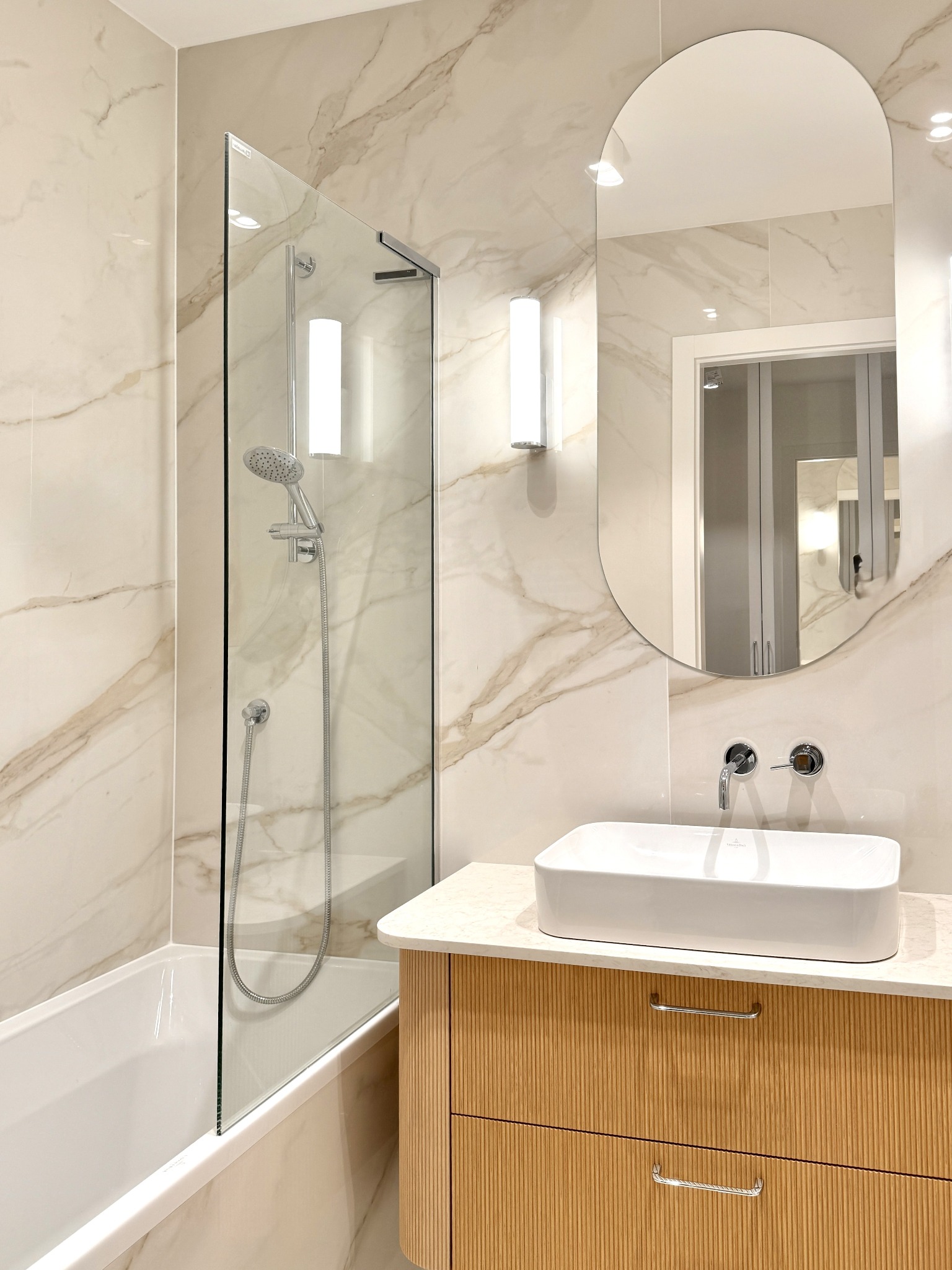 Nowoczesna łazienka z marmurowymi płytkami, owalnym lustrem, białą prostokątną umywalką nablatową i kabiną prysznicową ze szklaną ścianką.
