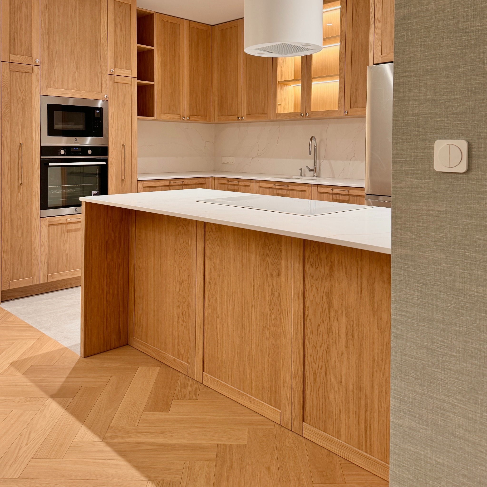 Jasna kuchnia z drewnianymi szafkami, białym blatem i parkietem w jodełkę, widok na wyspę kuchenną i zabudowane sprzęty.