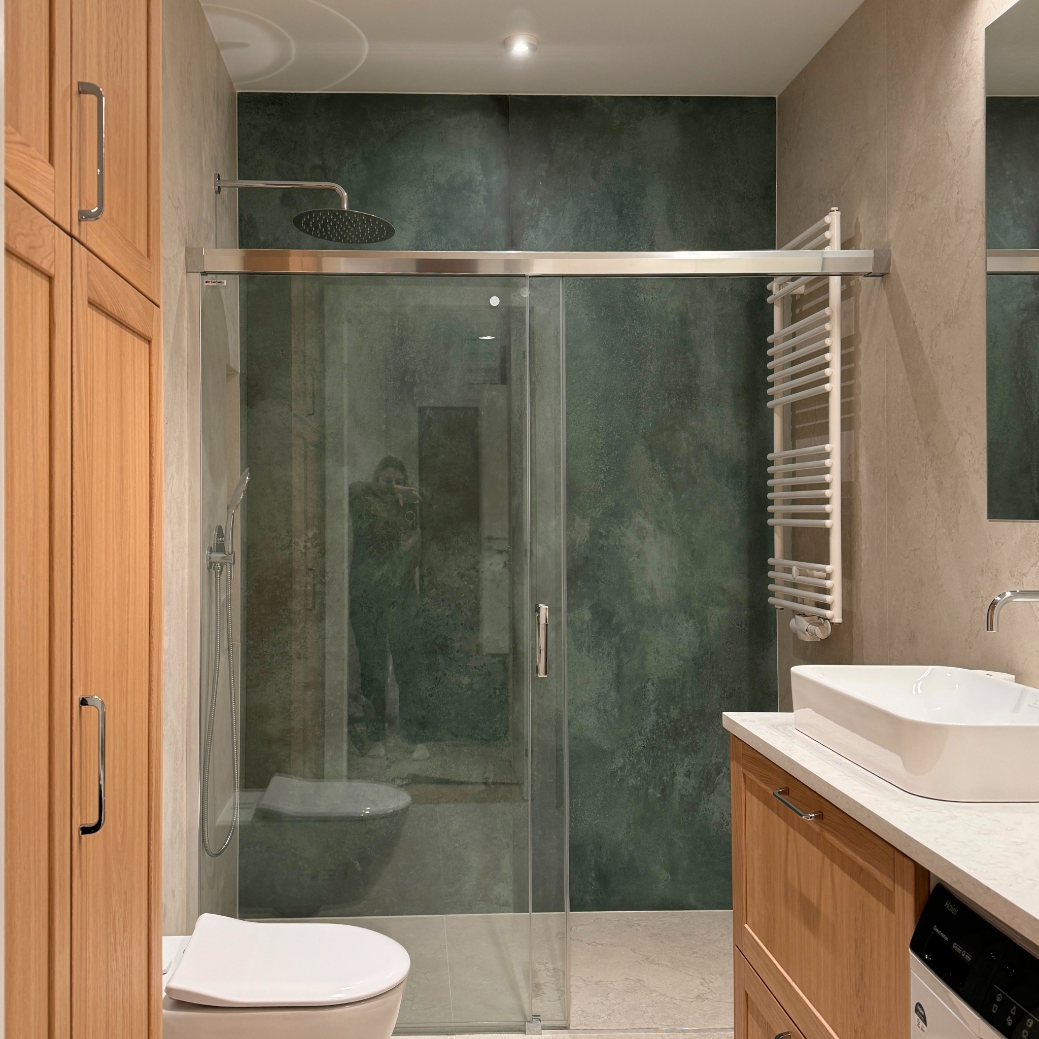 Nowoczesna łazienka z kabiną prysznicową typu walk-in, zieloną ścianą imitującą kamień, drewnianymi szafkami i umywalką nablatową; w szkle kabiny odbija się osoba robiąca zdjęcie.