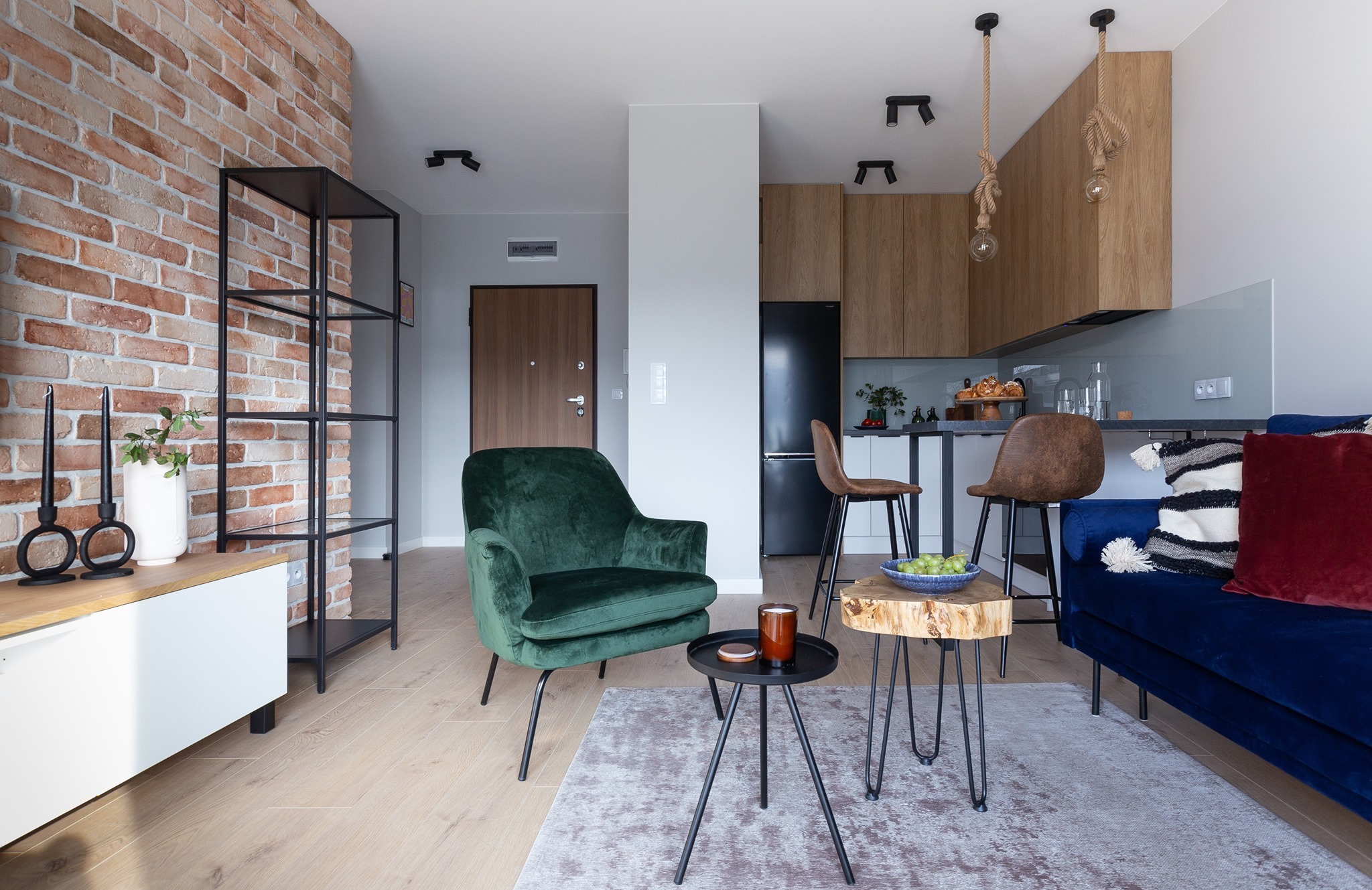 Salon z ceglaną ścianą, zielonym fotelem, niebieską sofą i elementami drewnianymi, oświetlony światłem dziennym i sztucznym.