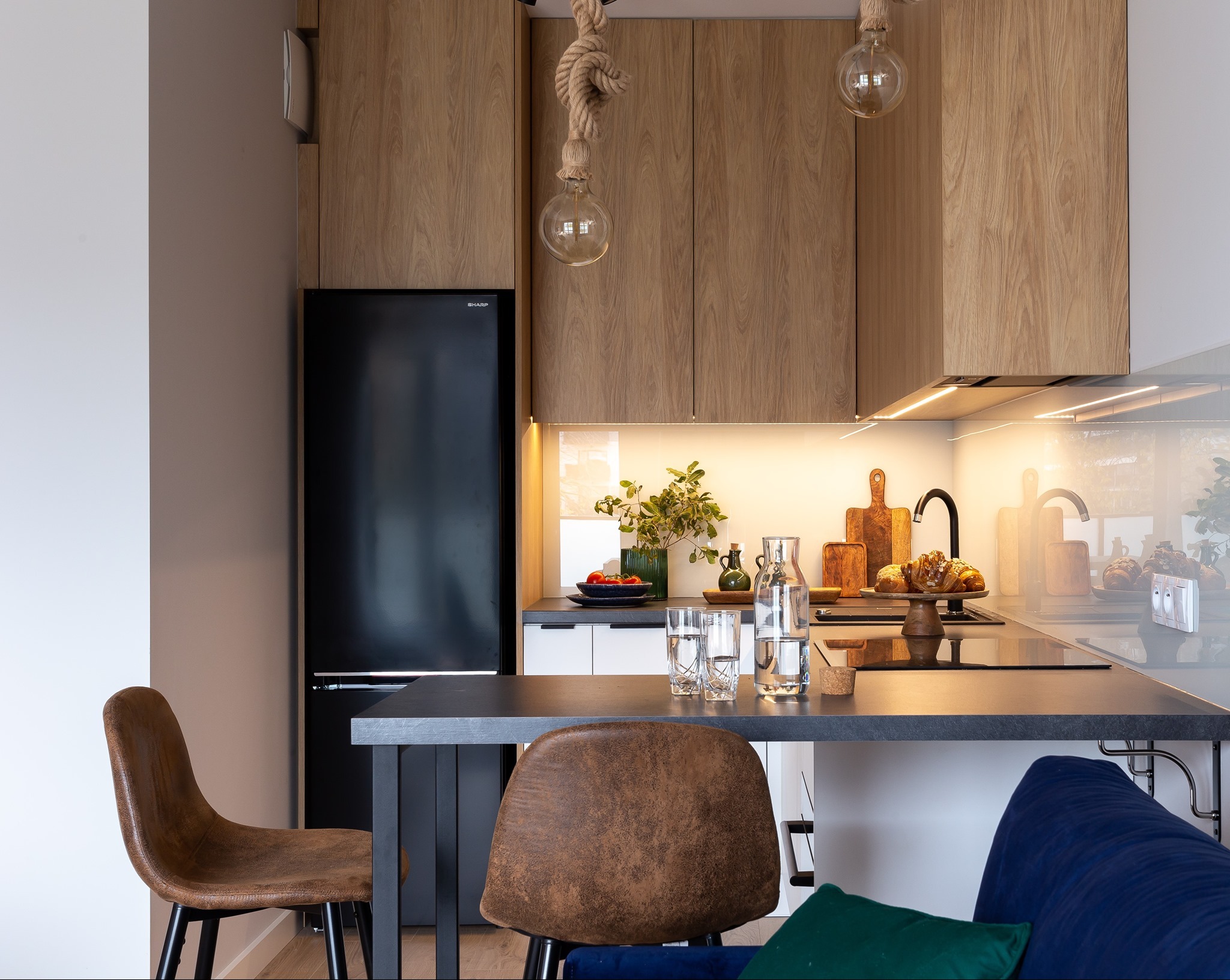 Nowoczesna kuchnia z drewnianymi szafkami, czarną lodówką Sharp, blatem z szarego kamienia i dekoracyjnymi żarówkami na sznurze, uzupełniona brązowymi krzesłami i roślinami.