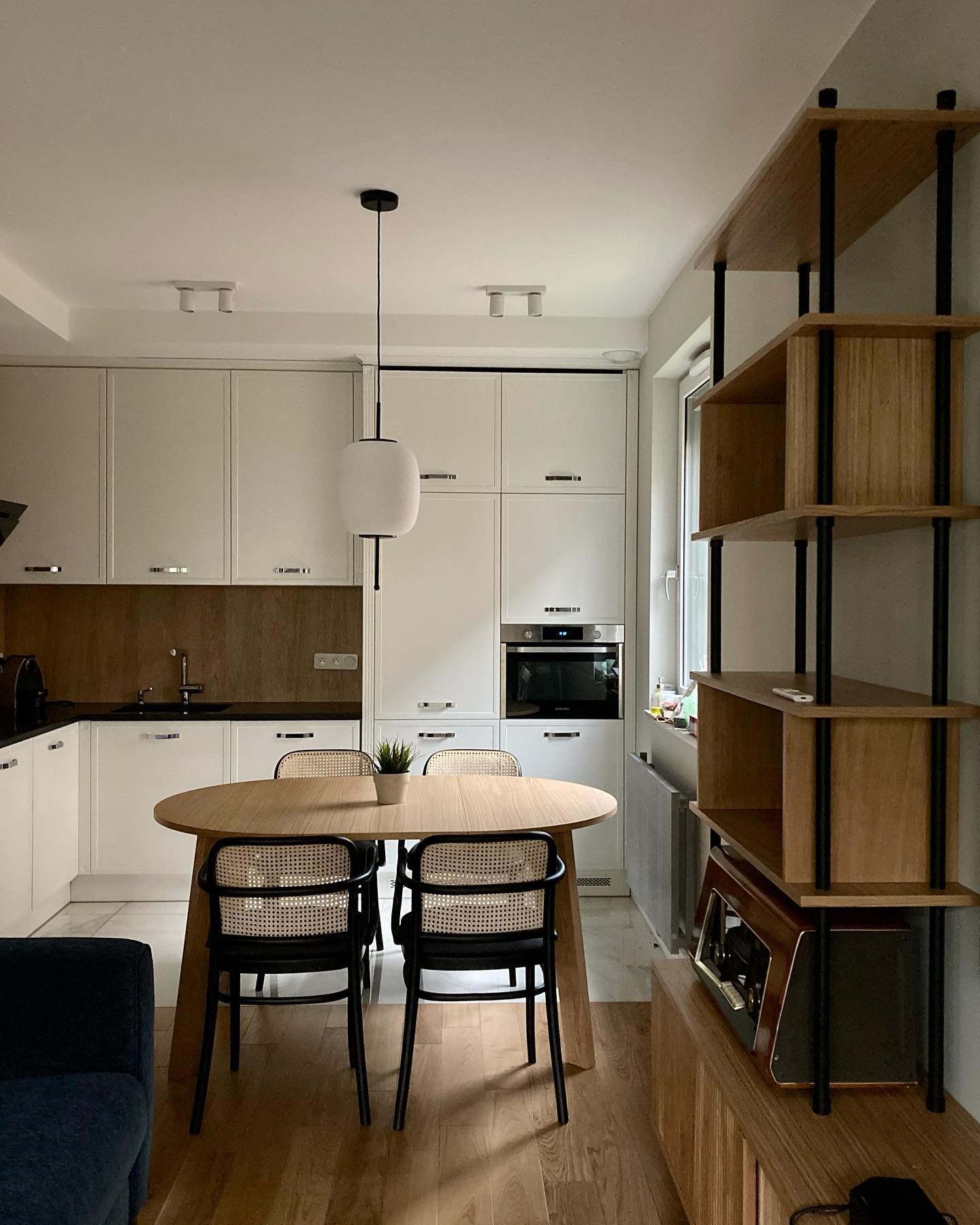 Jasna kuchnia z drewnianym stołem i krzesłami z rattanowym oparciem, uzupełniona regałem z drewnianymi półkami i czarnymi wspornikami oraz wiszącą lampą z mlecznym kloszem.