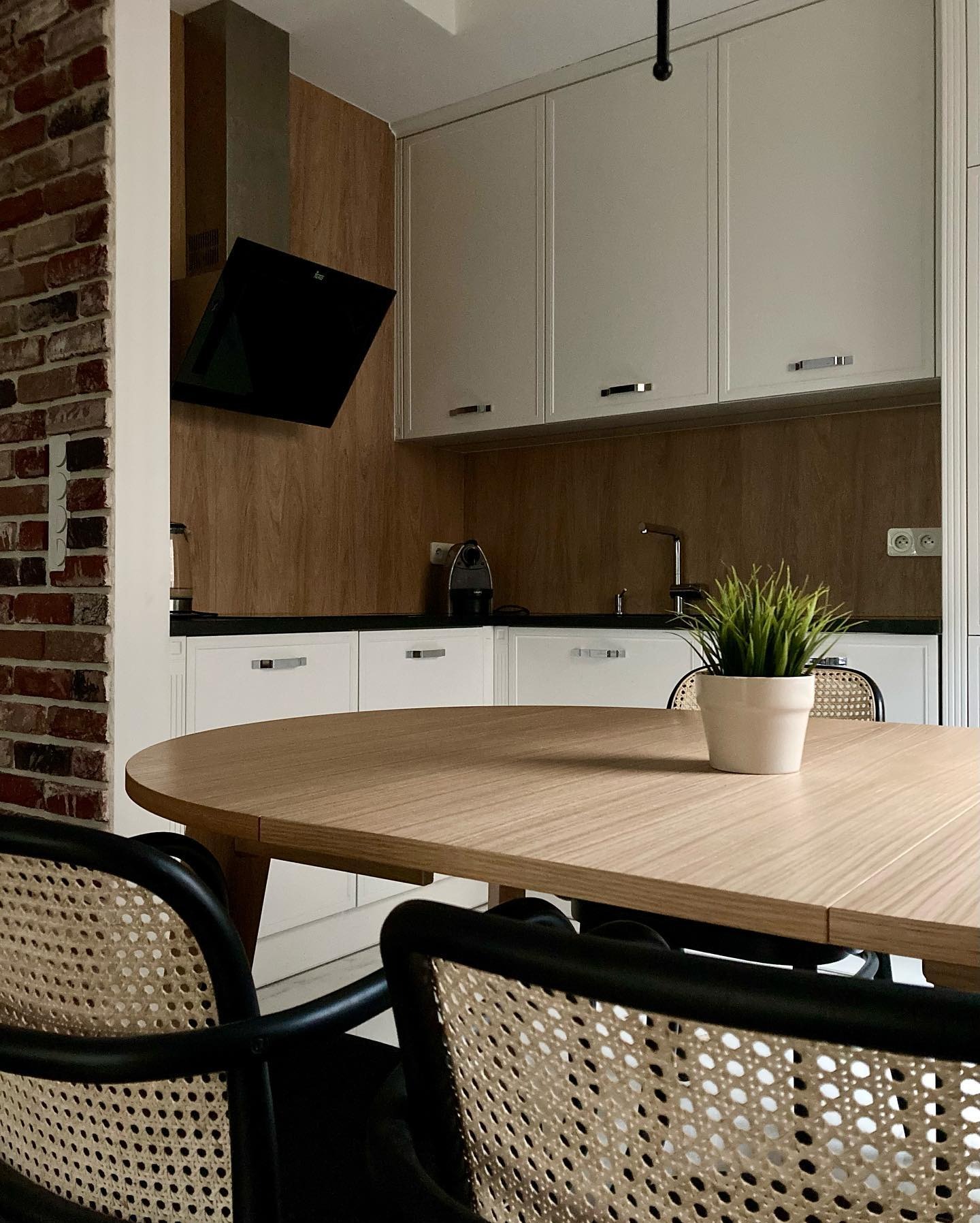 Jasna kuchnia z elementami cegły, okrągły drewniany stół z rośliną w doniczce, krzesła z rattanowym oparciem, czarny okap nad blatem.