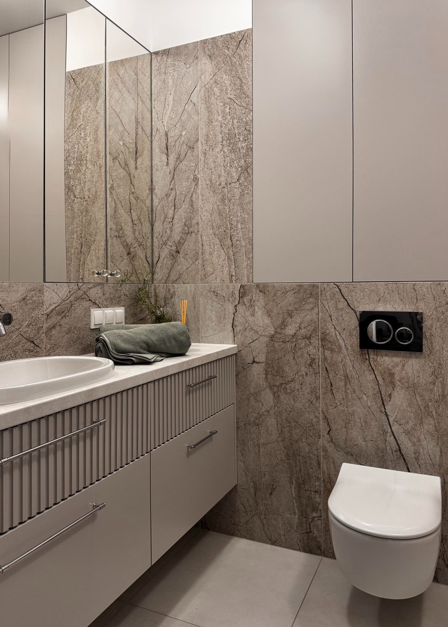 Elegancka łazienka w neutralnych barwach: umywalka nablatowa, marmurowe płytki, podwieszana toaleta i minimalistyczne szafki. Całość dopełnia tekstylny akcent.