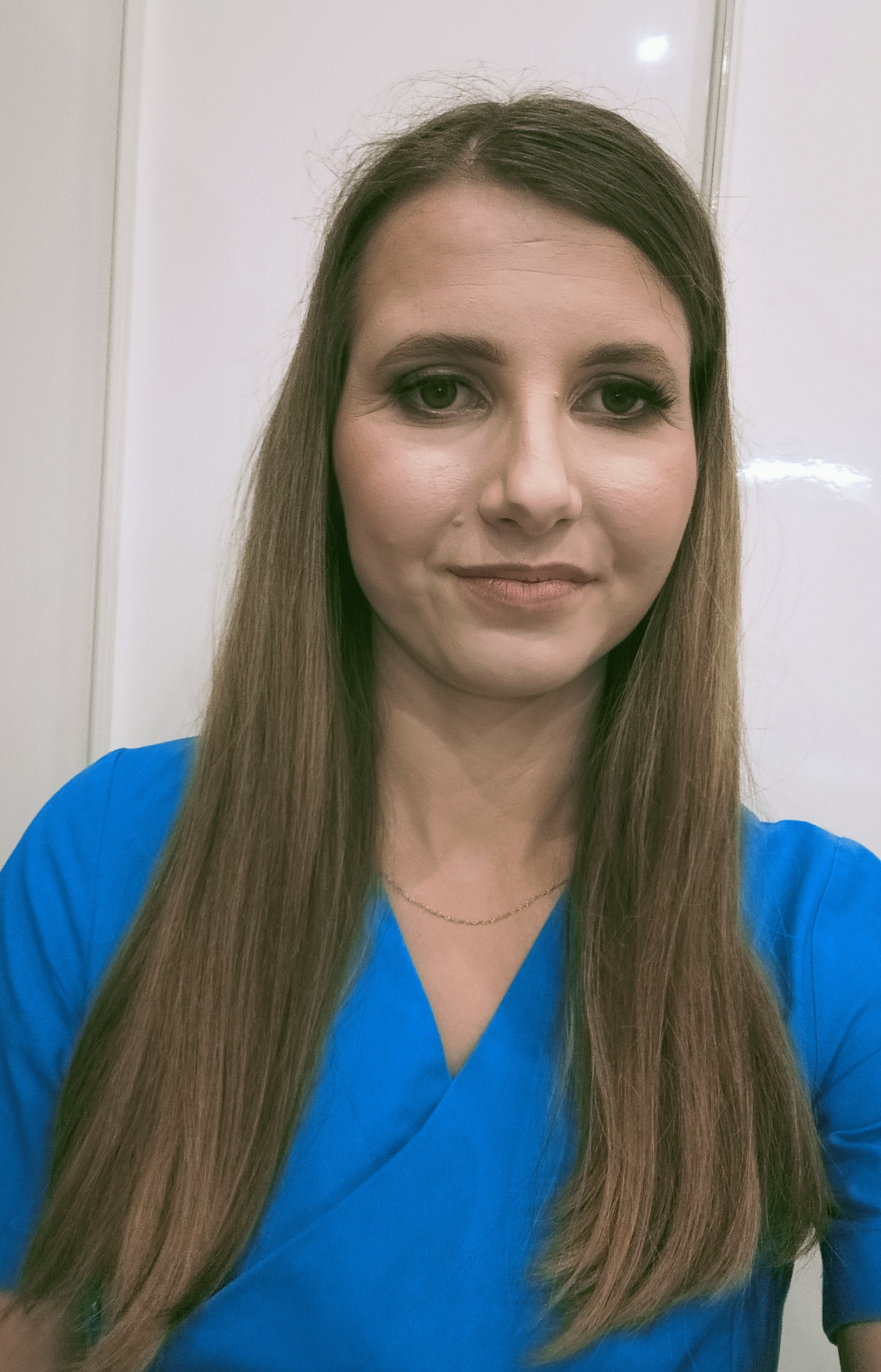 Portret kobiety w niebieskim uniformie, z długimi brązowymi włosami i delikatnym makijażem, stojącej na tle białej ściany.