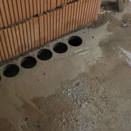 JW-WENTAR - Równy rząd okrągłych otworów wywierconych w mokrym, szarym betonie przy ścianie z cegieł ceramicznych.