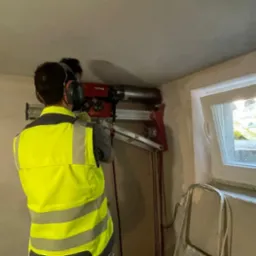 Pracownik w kamizelce odblaskowej wierci wiertnicą koronową otwór w ścianie nad oknem, widoczne przewody chłodzące i drabina oparta o ścianę.