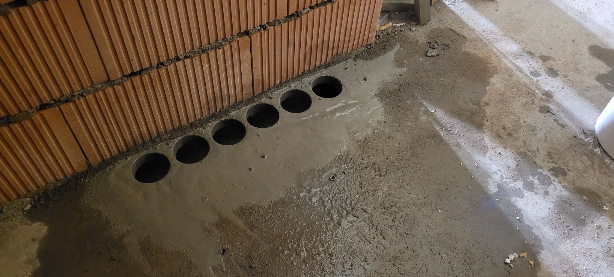 Równy rząd okrągłych otworów wywierconych w mokrym, szarym betonie przy ścianie z cegieł ceramicznych.
