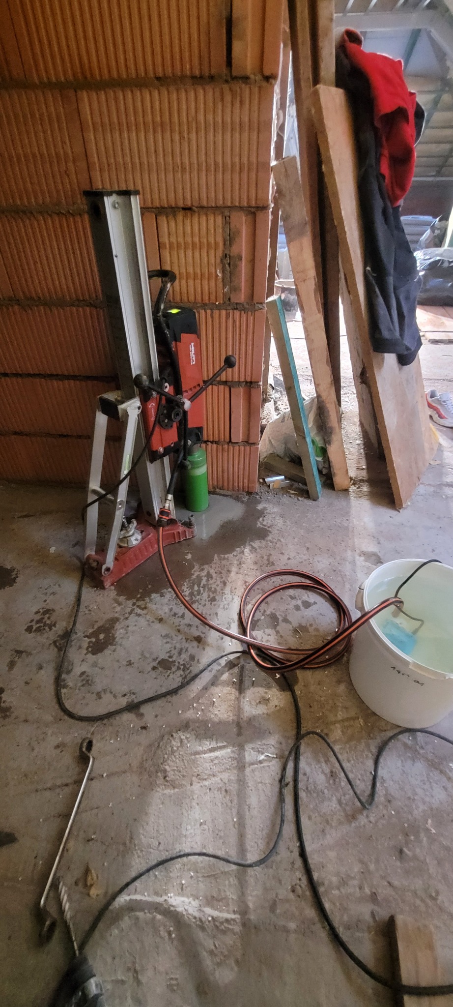 Czerwona wiertnica rdzeniowa Hilti, ustawiona na statywie przed ścianą z cegieł dziurawki, podłączona do wiadra z wodą; widoczne przewody i wąż doprowadzający wodę.