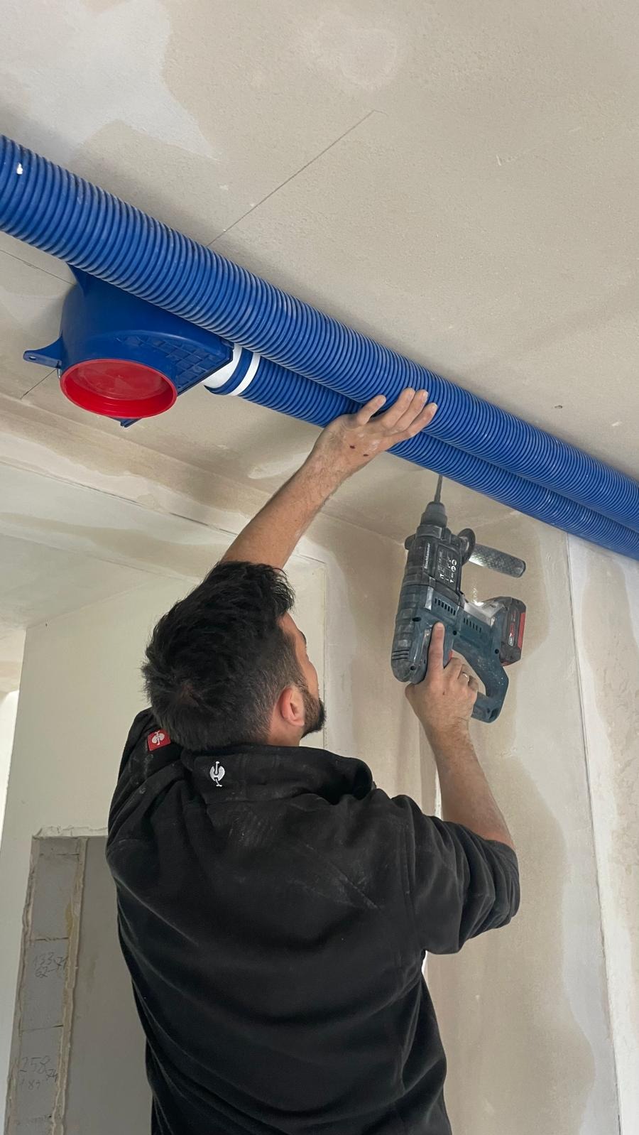 Pracownik montuje niebieskie rury wentylacyjne pod sufitem za pomocą wiertarki udarowej, widoczny fragment instalacji z czerwonym elementem.
