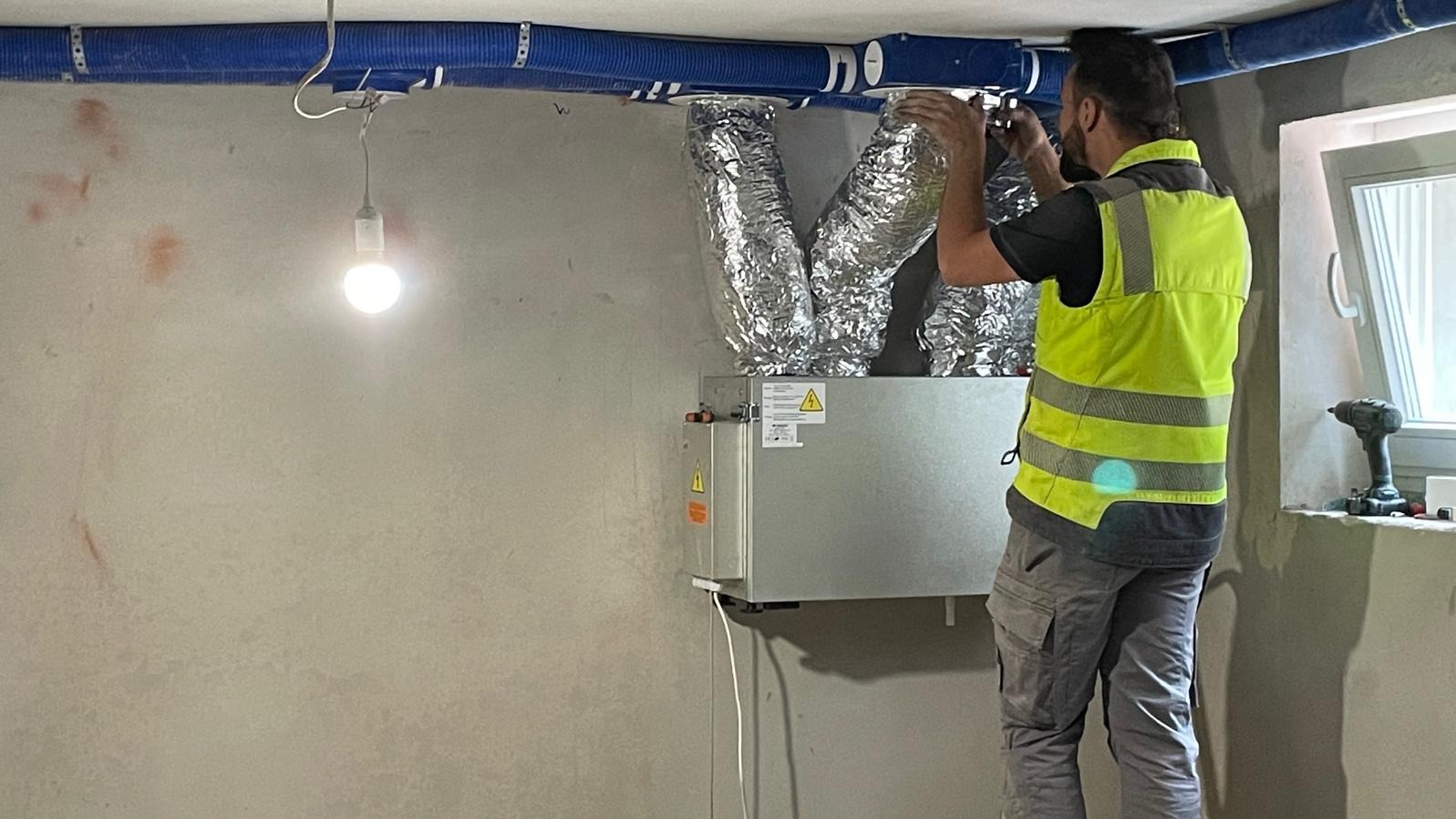 Instalator w kamizelce odblaskowej podłącza aluminiowe rury wentylacyjne do centrali wentylacyjnej w surowym wnętrzu, widoczne niebieskie rury wentylacyjne pod sufitem i wisząca żarówka.