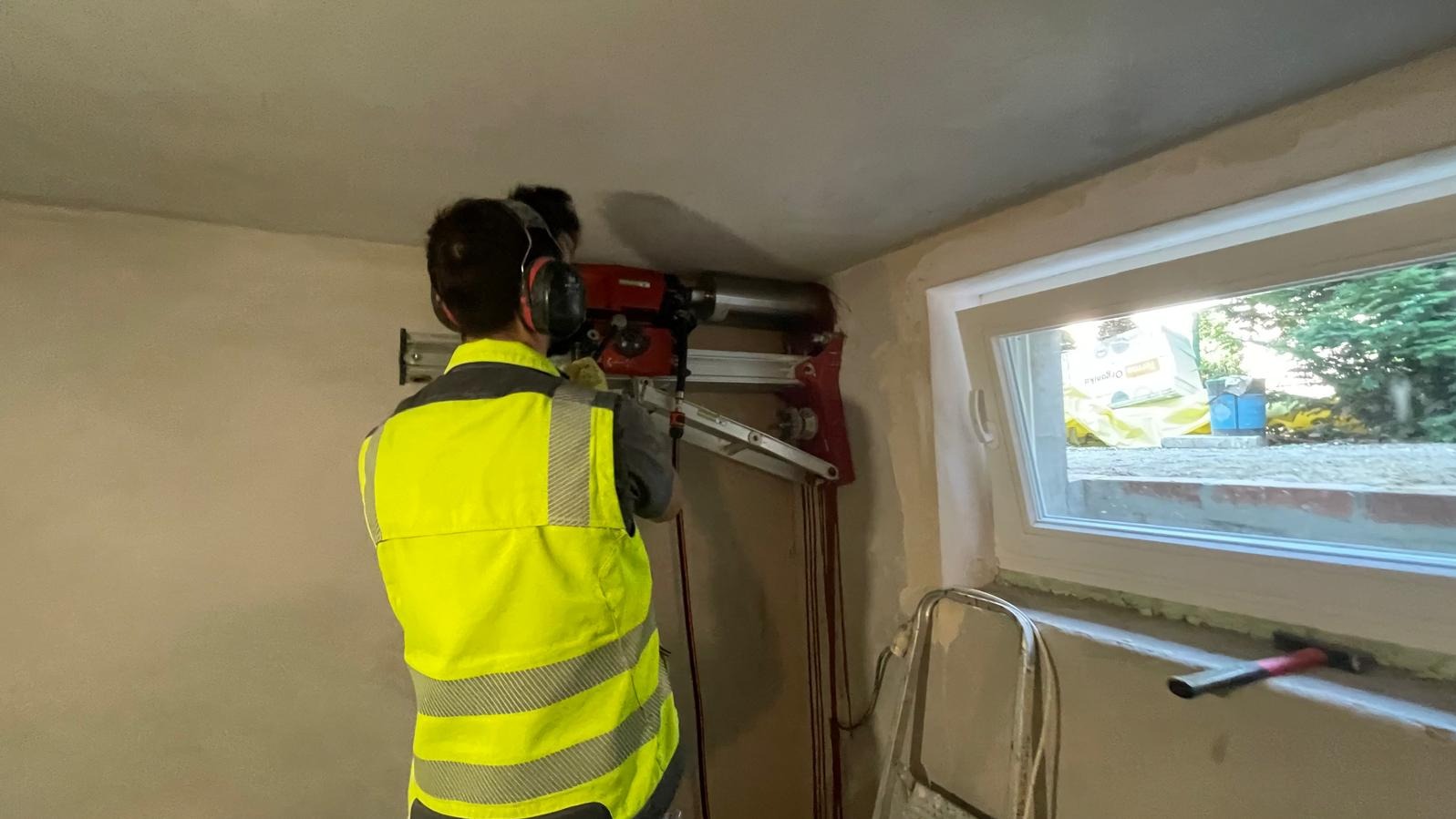 Pracownik w kamizelce odblaskowej wierci wiertnicą koronową otwór w ścianie nad oknem, widoczne przewody chłodzące i drabina oparta o ścianę.
