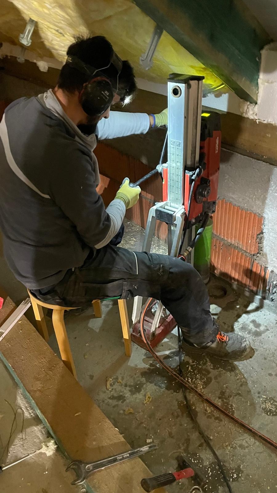 Dwóch mężczyzn w kaskach i słuchawkach ochronnych, wiercących otwór w ceglanej ścianie za pomocą wiertnicy Hilti, siedząc na niskim stołku w pomieszczeniu z drewnianą konstrukcją stropu.