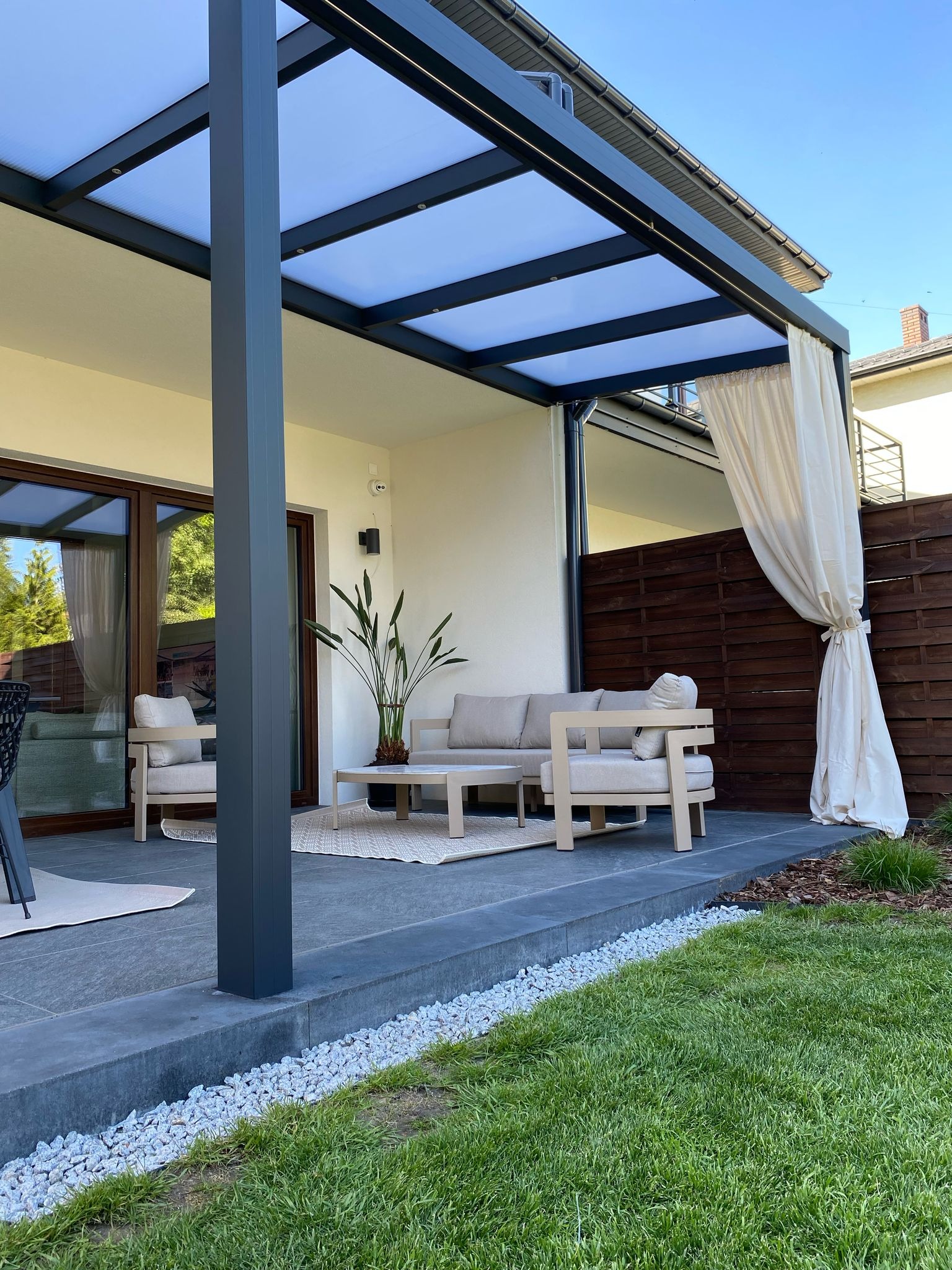 Nowoczesna pergola metalowa z oświetleniem profilowym, jasne meble ogrodowe, zasłona i zadbany trawnik tworzą przytulną strefę relaksu na tarasie.