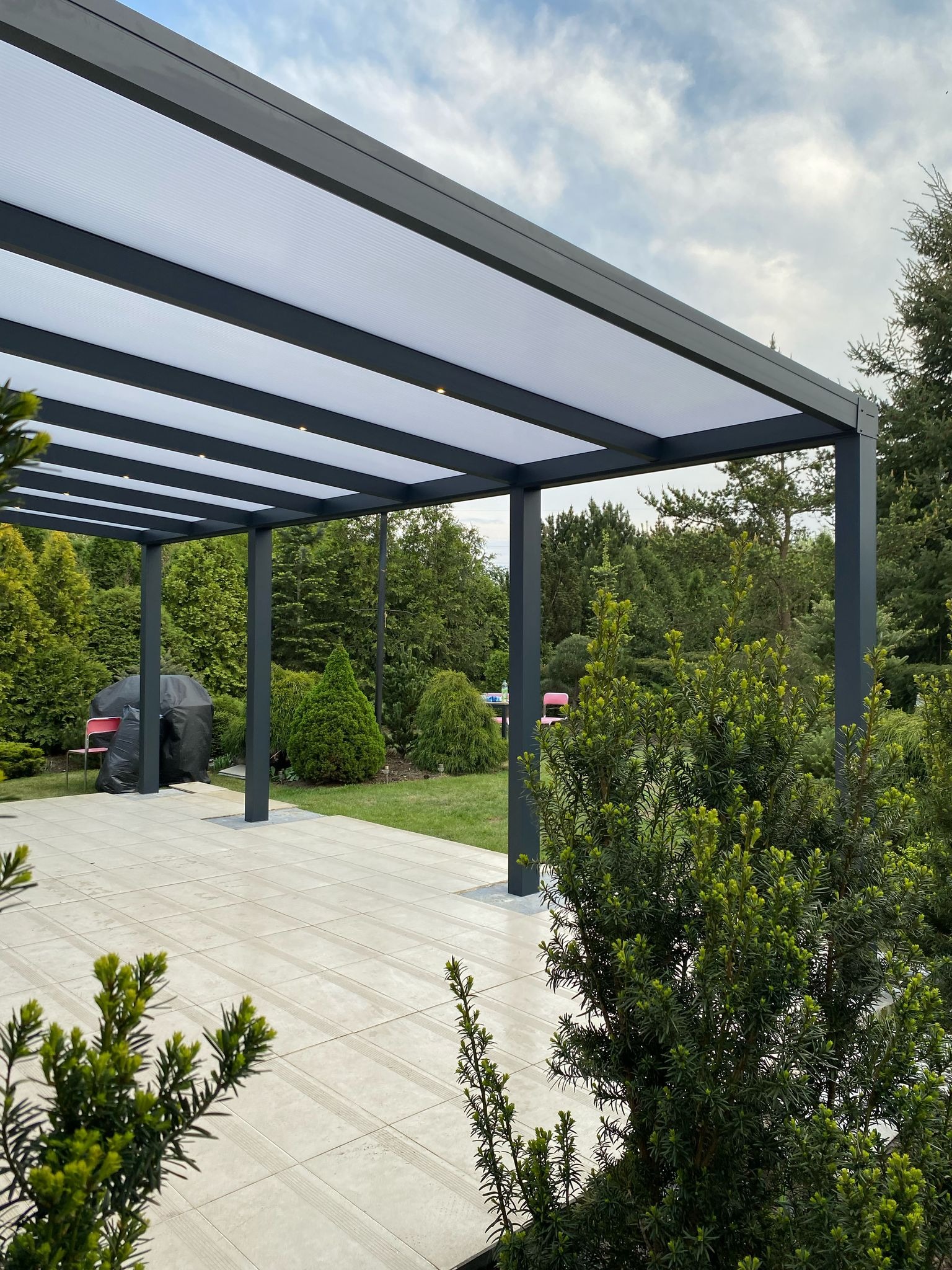 Nowoczesna, szara pergola metalowa z oświetleniem punktowym w profilach, na tle zieleni i błękitnego nieba. Widok na taras z płyt.