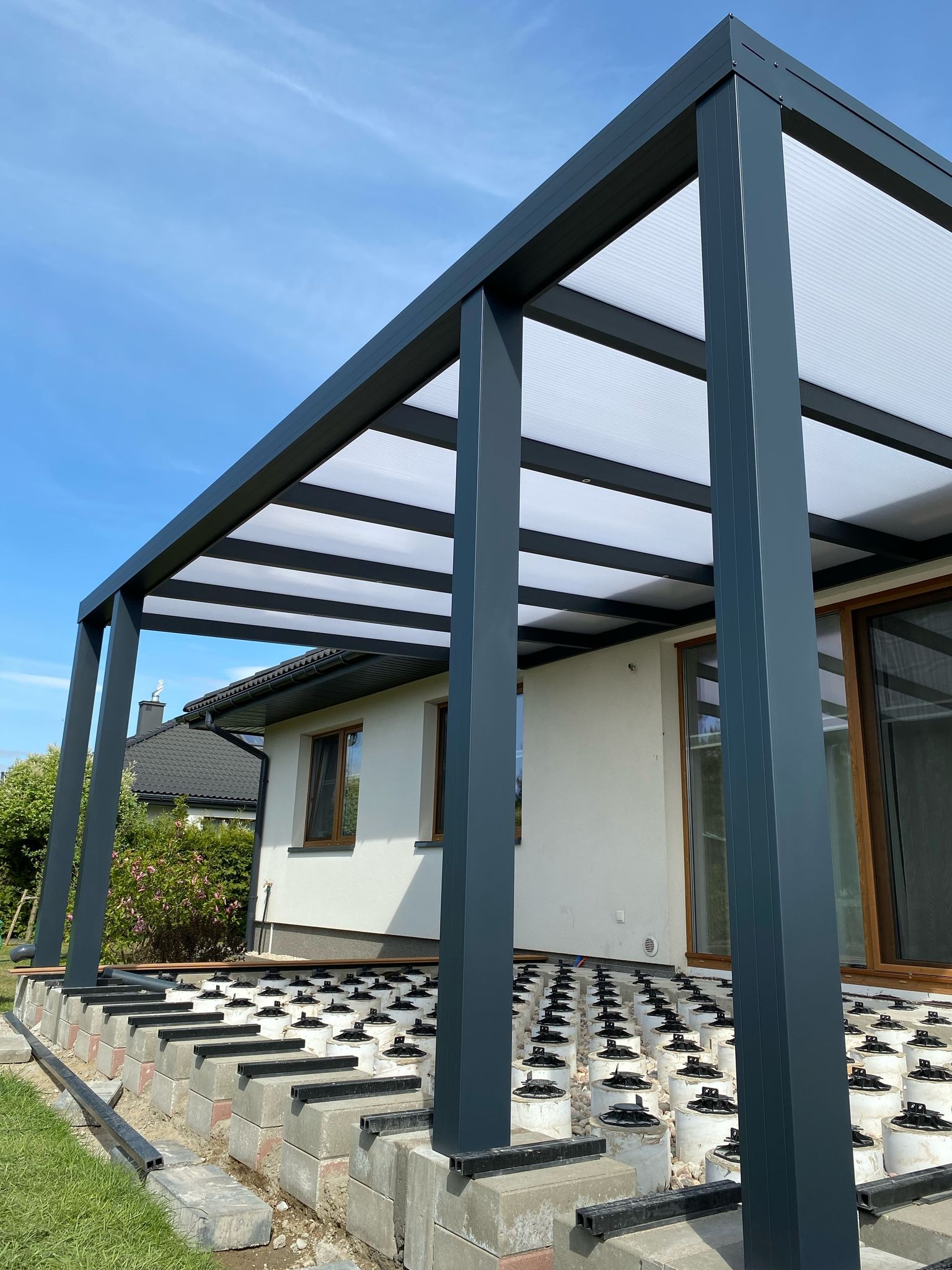 Montaż metalowej pergoli z mlecznym zadaszeniem na tarasie. Widoczne betonowe podstawy pod konstrukcję i elementy oświetlenia w profilach.