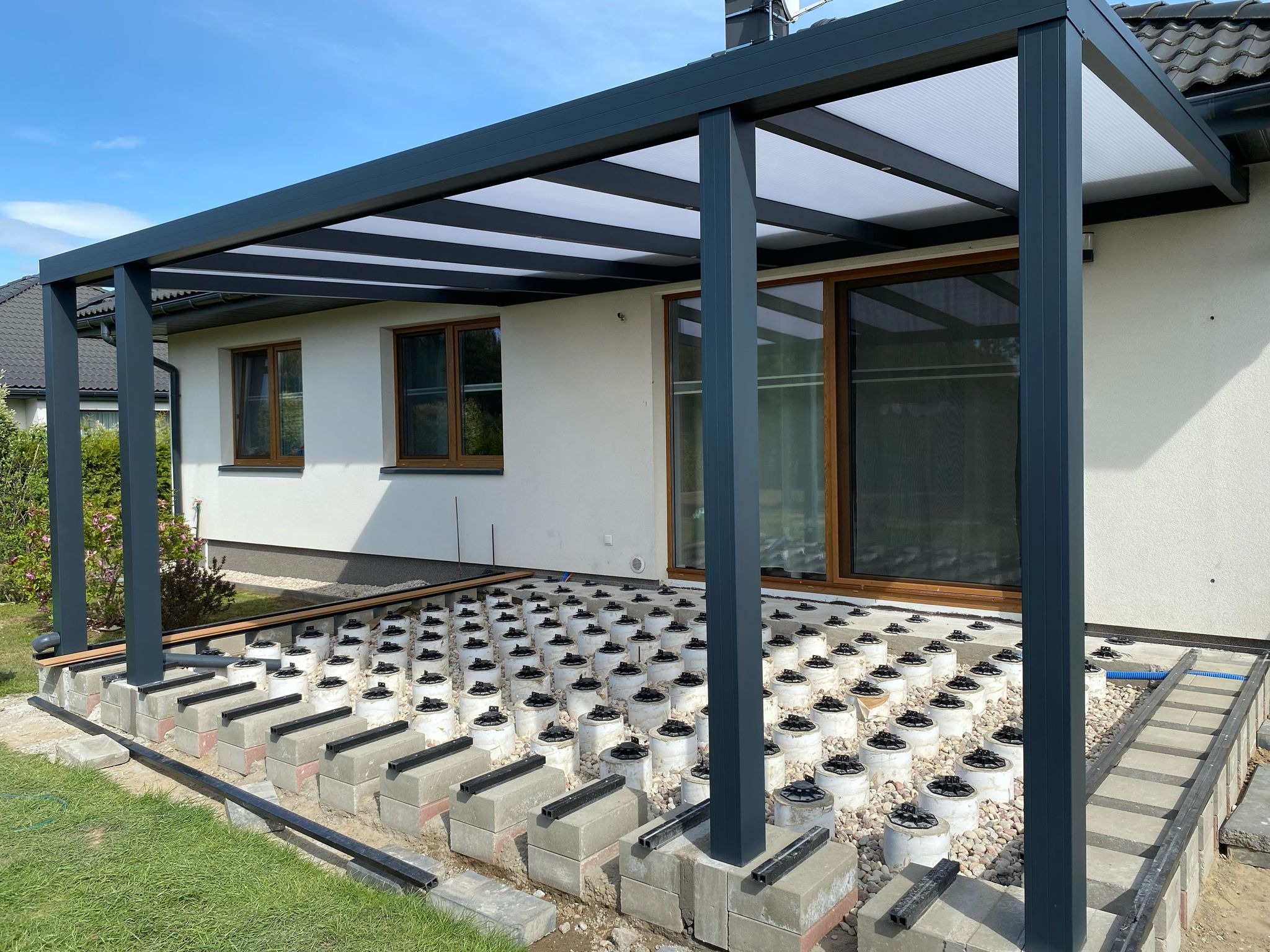 Montaż metalowej pergoli przy białym domu. Widoczne podpory tarasowe i fragment konstrukcji z oświetleniem w profilach. Błękitne niebo w tle.