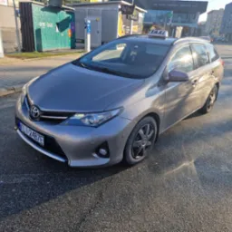 Szary samochód typu kombi z napisem TAXI na dachu, zaparkowany na ulicy w słoneczny dzień, widoczny automat paczkowy DPD i budynek z reklamą LVBet w tle.