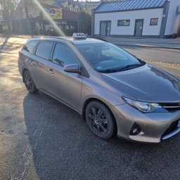 GREGOR TAXI nr. 17 - Szary samochód kombi z napisem TAXI na dachu, zaparkowany na ulicy w słoneczny dzień, z widocznymi budynkami w tle.